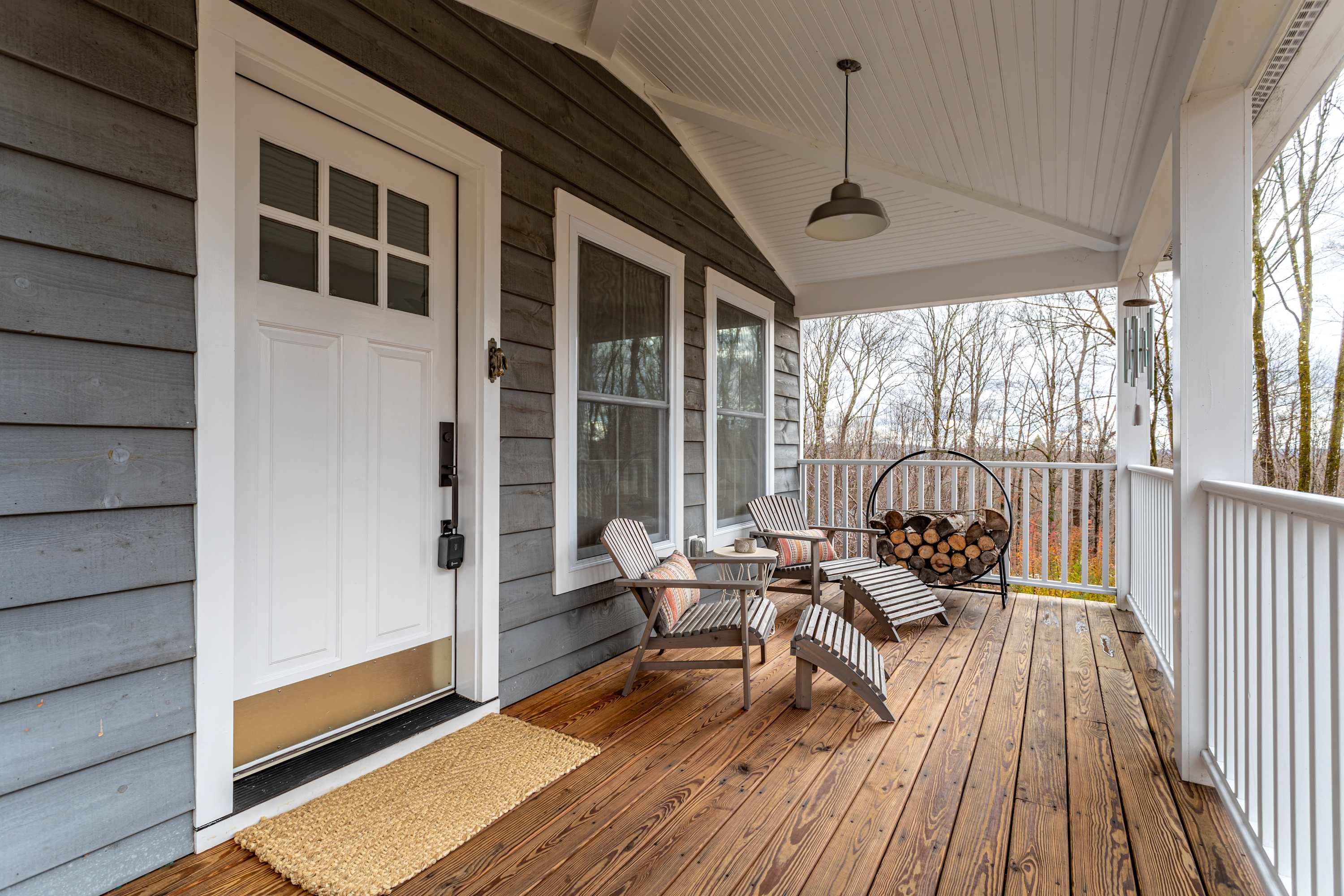 A welcoming front porch features a white entry door, gray siding, and a wooden floor, furnished with two wooden lounge chairs and a log holder filled with firewood. The porch offers a cozy spot to relax with views of the surrounding natural environment.