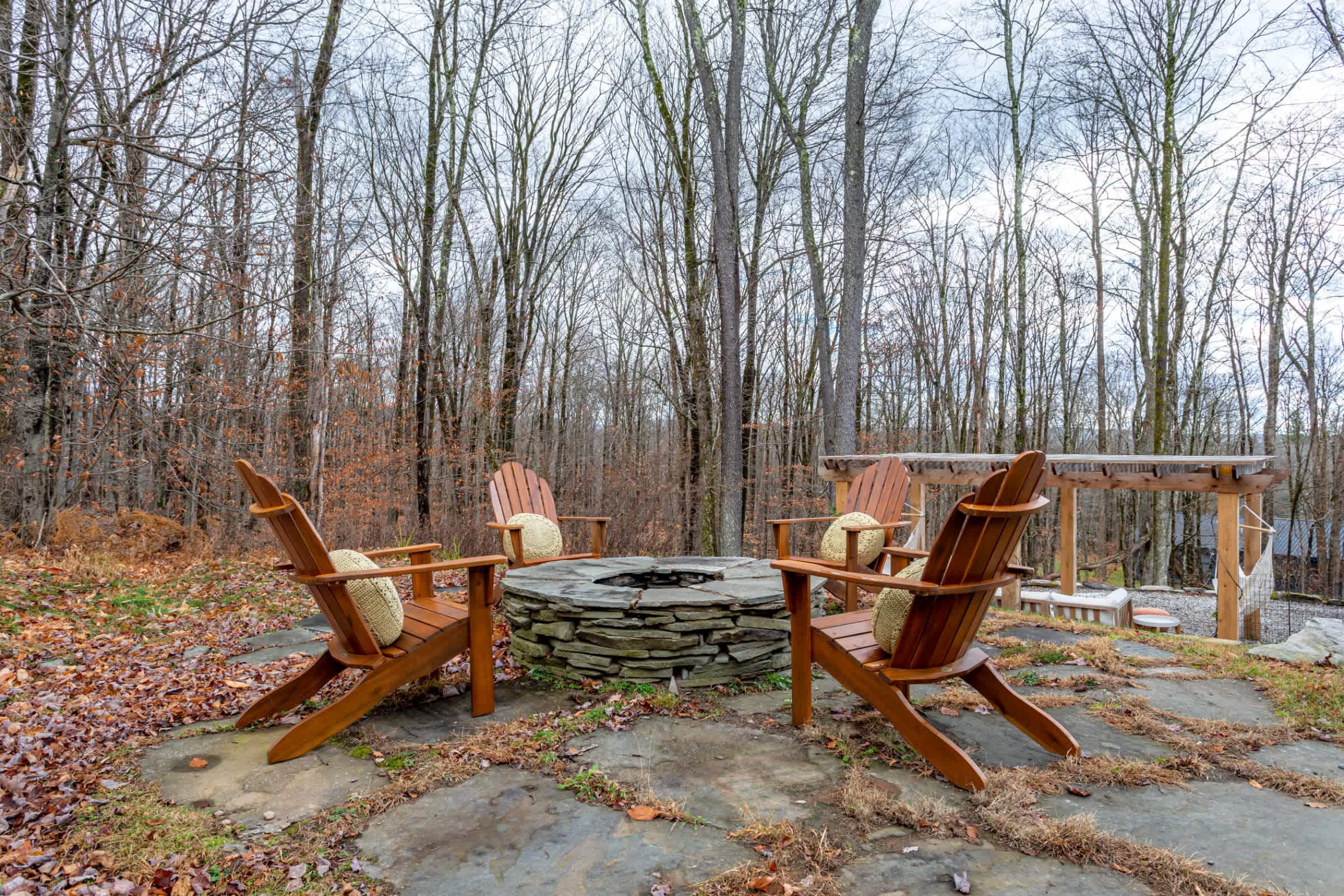 A stone-ringed fire pit is surrounded by four wooden Adirondack chairs with cushions, set on a flagstone patio amidst fallen autumn leaves. Bare trees form a dense backdrop.