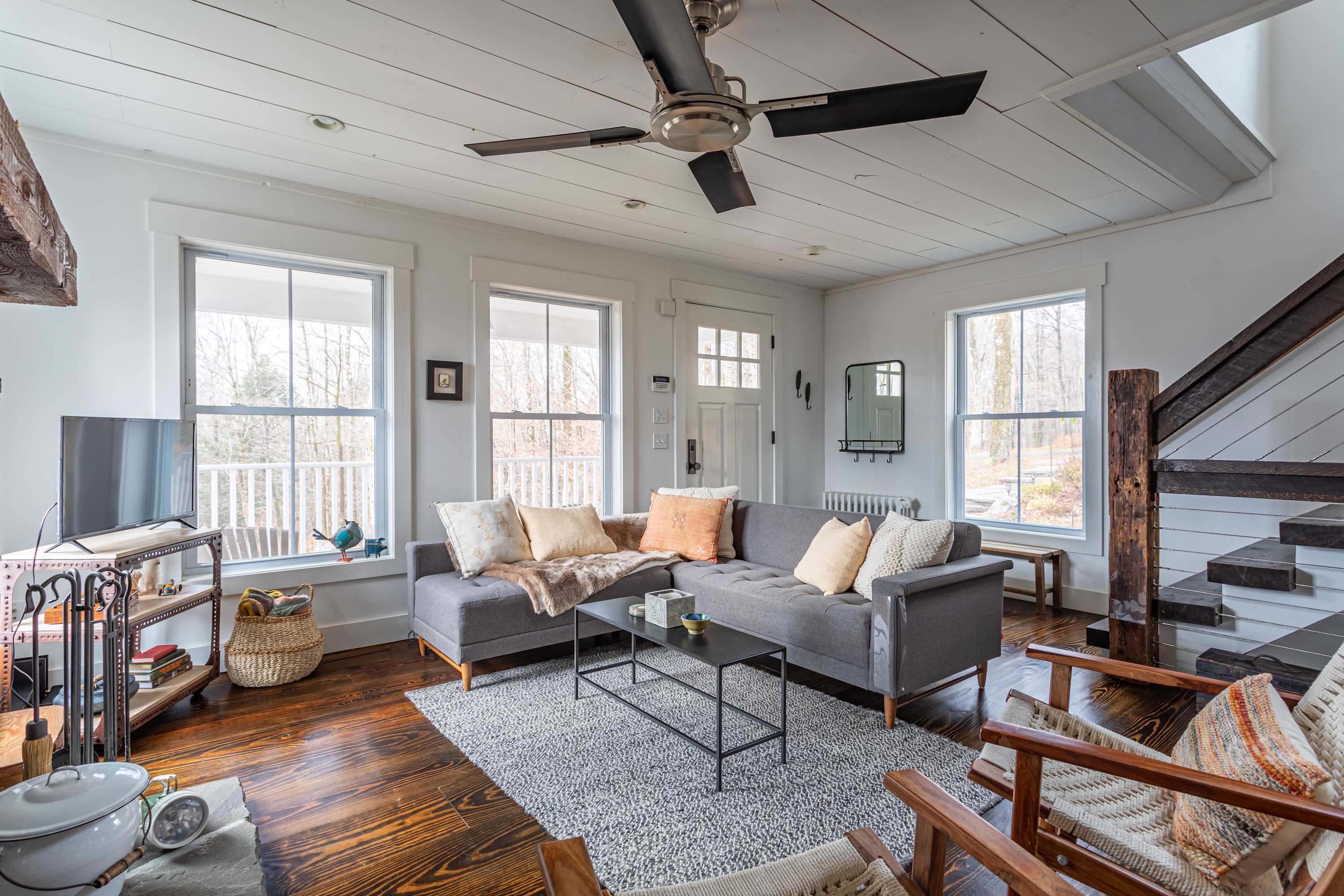 Bright living room featuring a grey sectional sofa, dark wood floors, a white shiplap ceiling with a ceiling fan, and a view towards the entrance and a rustic wooden staircase.