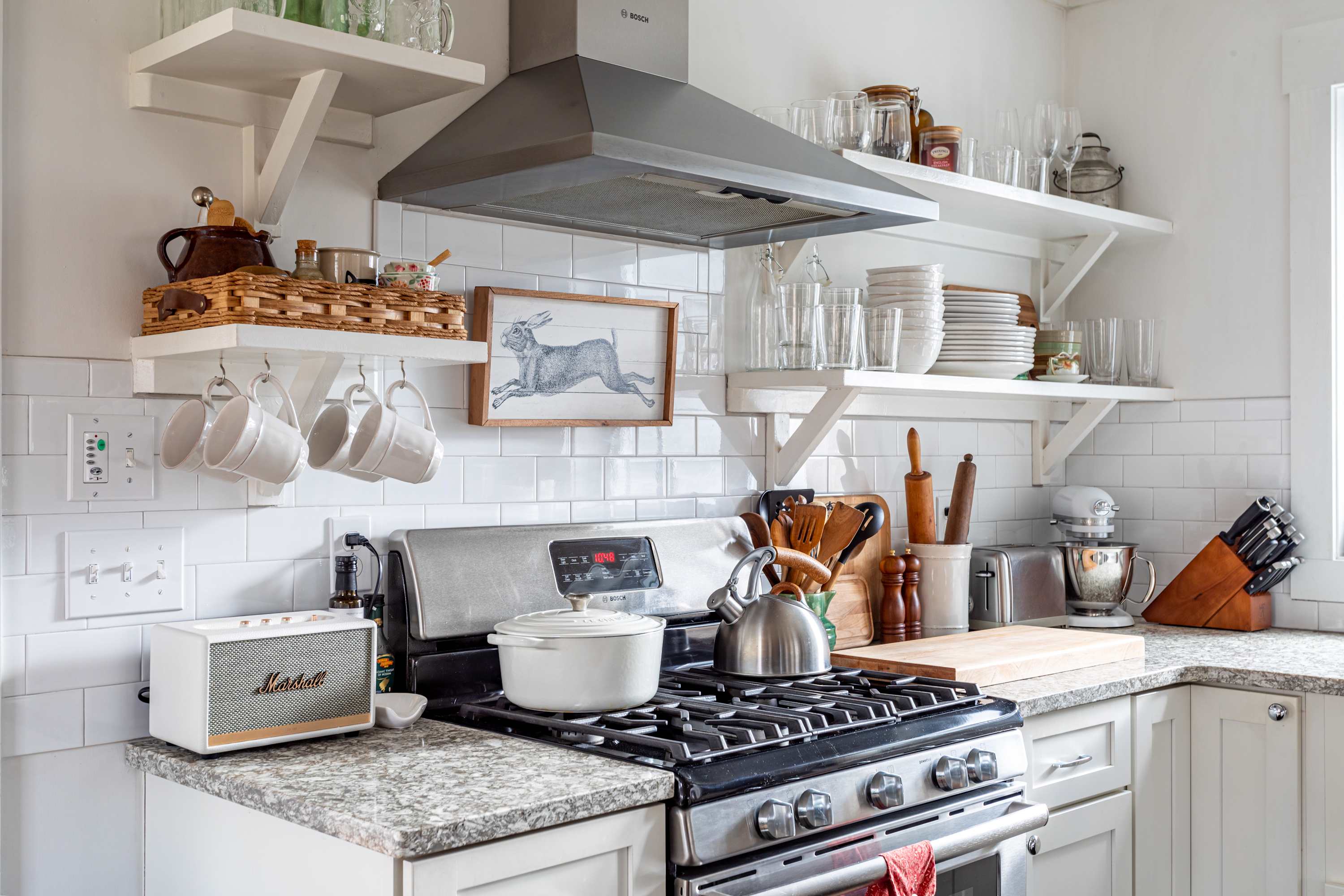 Modern farmhouse kitchen with white subway tile backsplash, open shelving, stainless steel gas range, and granite countertops, featuring a white cast iron pot on the stove.