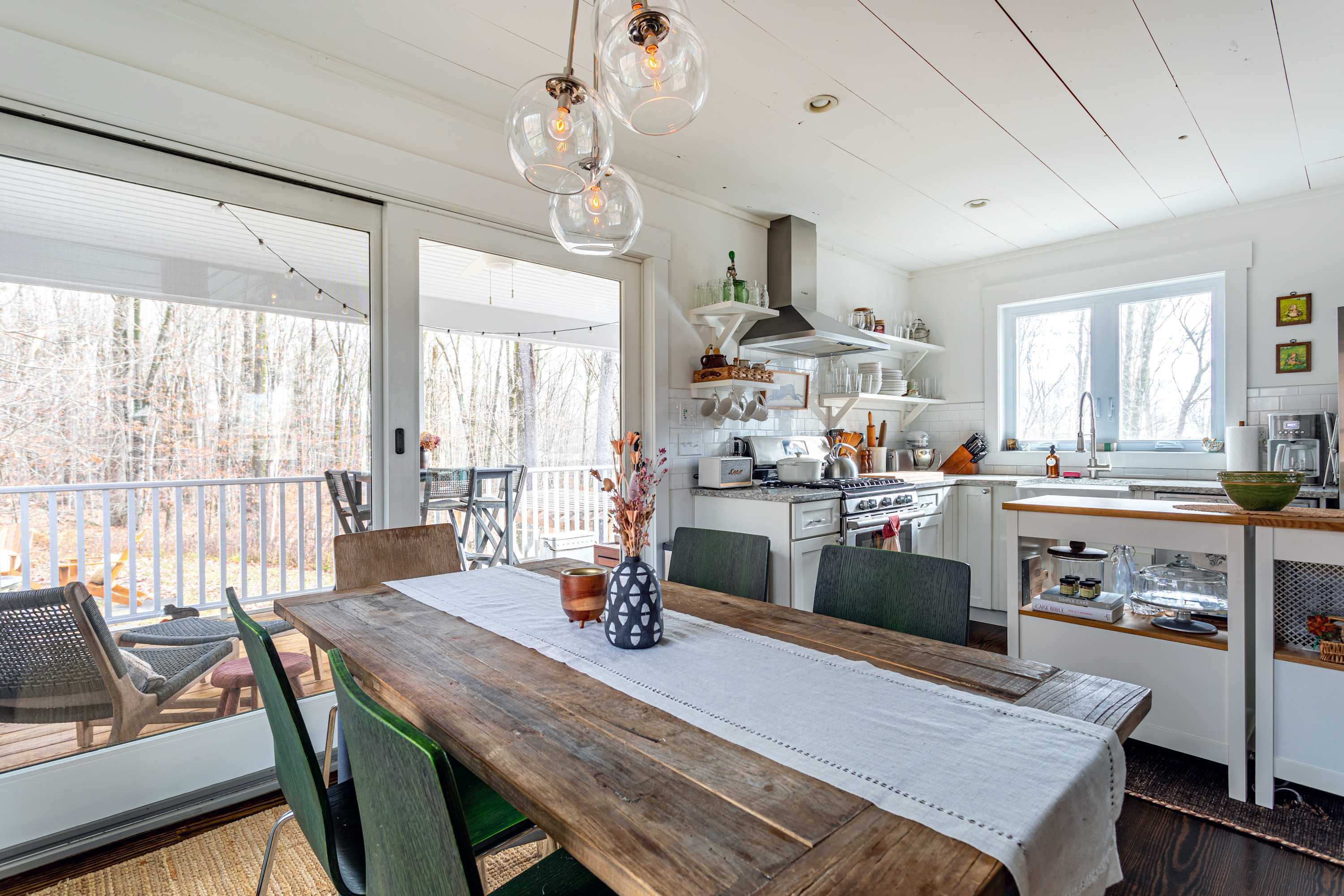 Rustic dining area with a long wooden table, mixed-style chairs, and a modern pendant light fixture, adjacent to the kitchen and featuring a large sliding glass door to an outdoor deck.