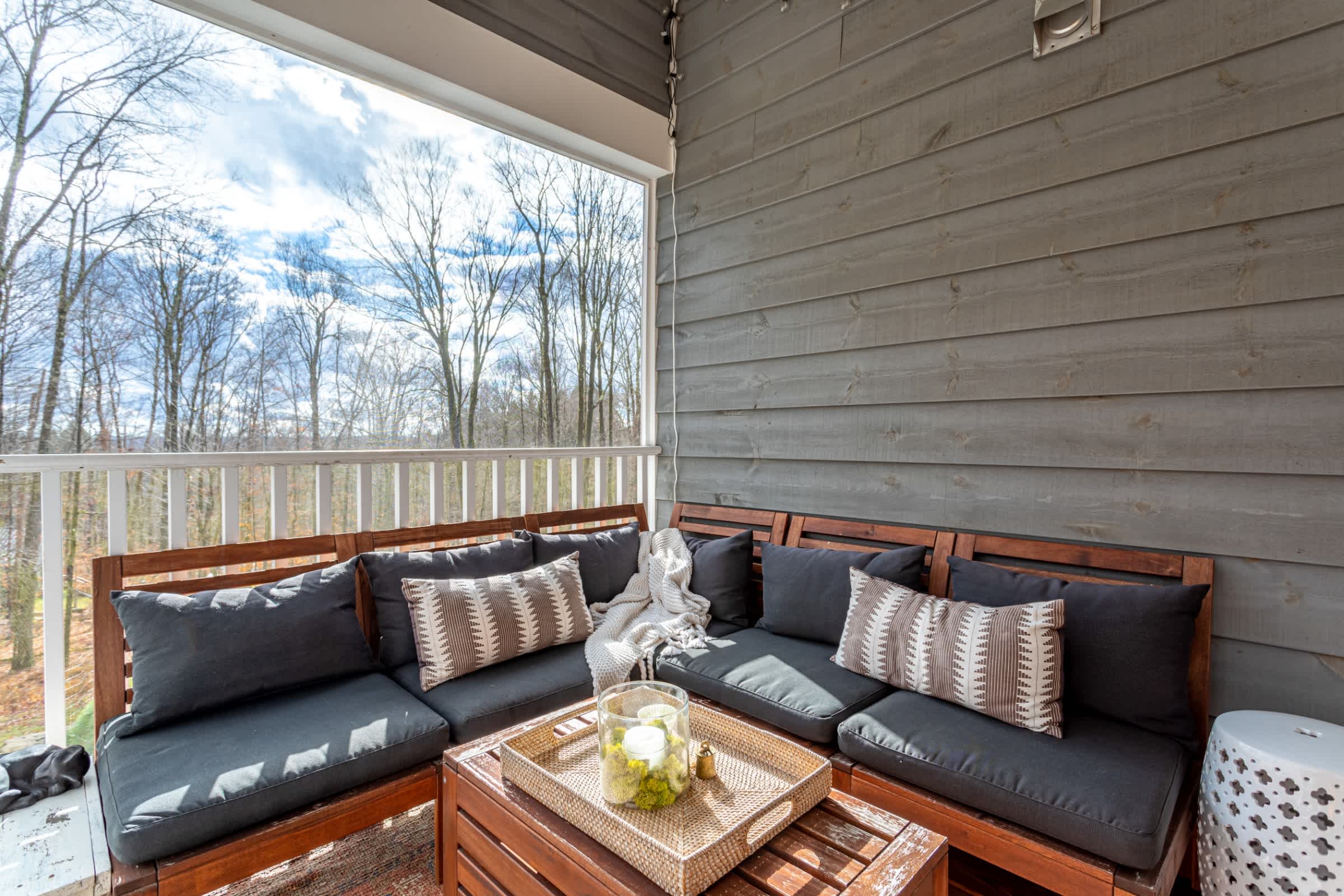 A cozy covered porch features a wooden slatted sofa with dark cushions and patterned throw pillows, centered around a slatted wooden coffee table with a decorative tray, overlooking a wooded landscape.