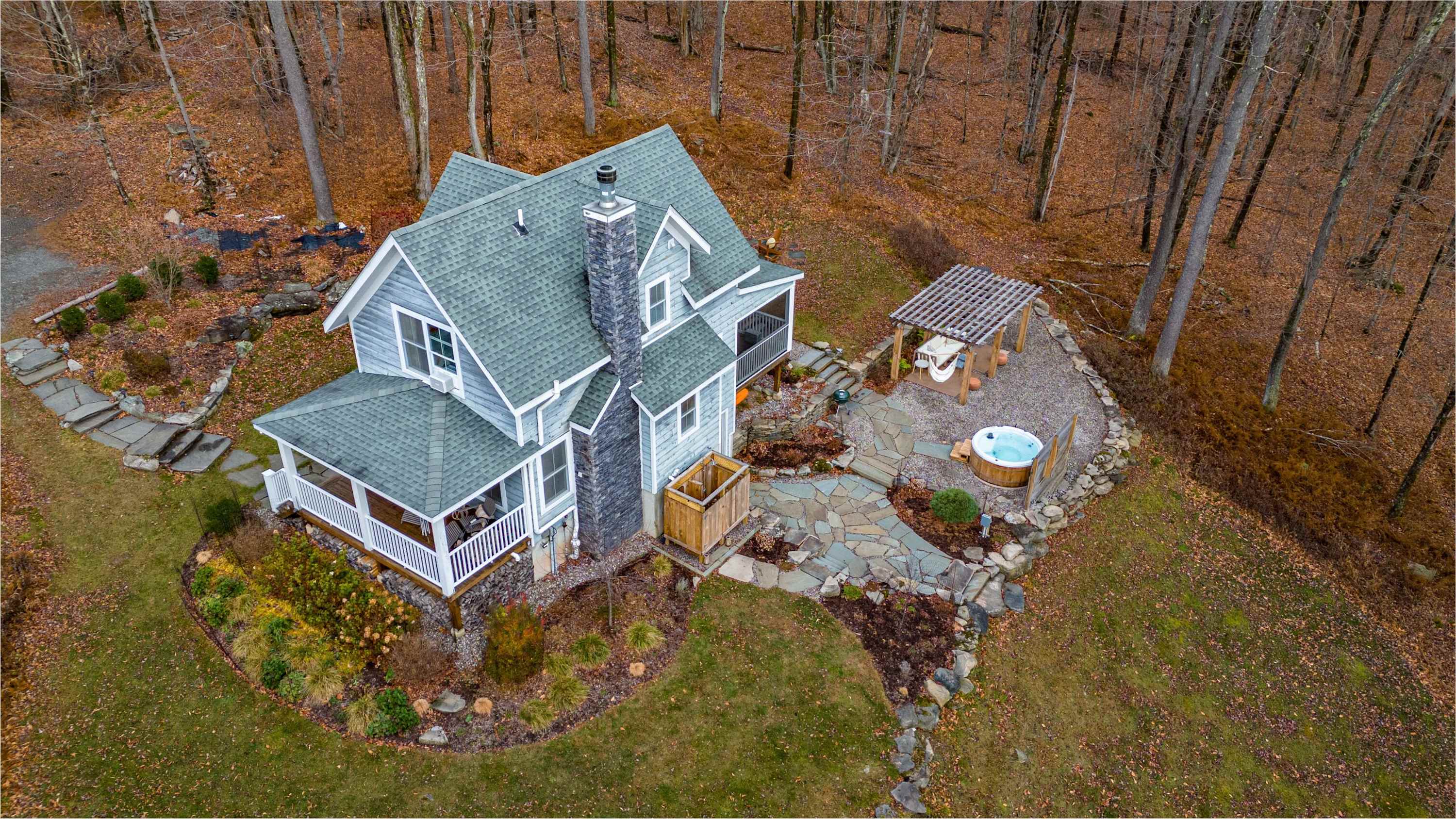 Aerial view of a light blue house with a dark green roof, nestled in a bare autumn forest, featuring a stone chimney, a hot tub, and a pergola on a gravel patio.