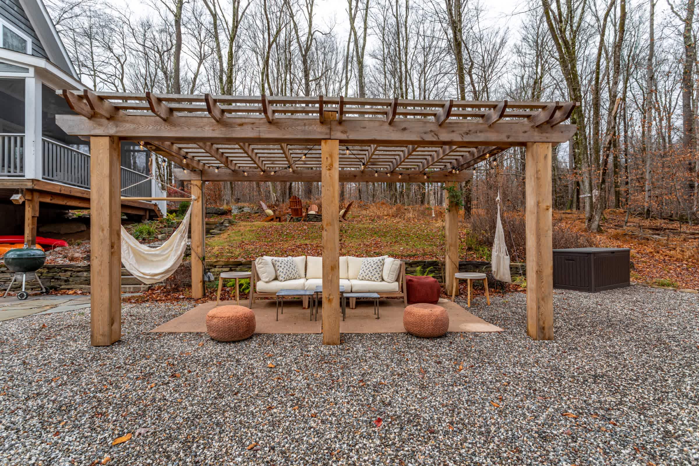 A spacious gravel patio features a large wooden pergola sheltering an outdoor living area with two white sofas, small tables, and orange poufs. Bare trees surround the area, and a hammock hangs from the pergola.