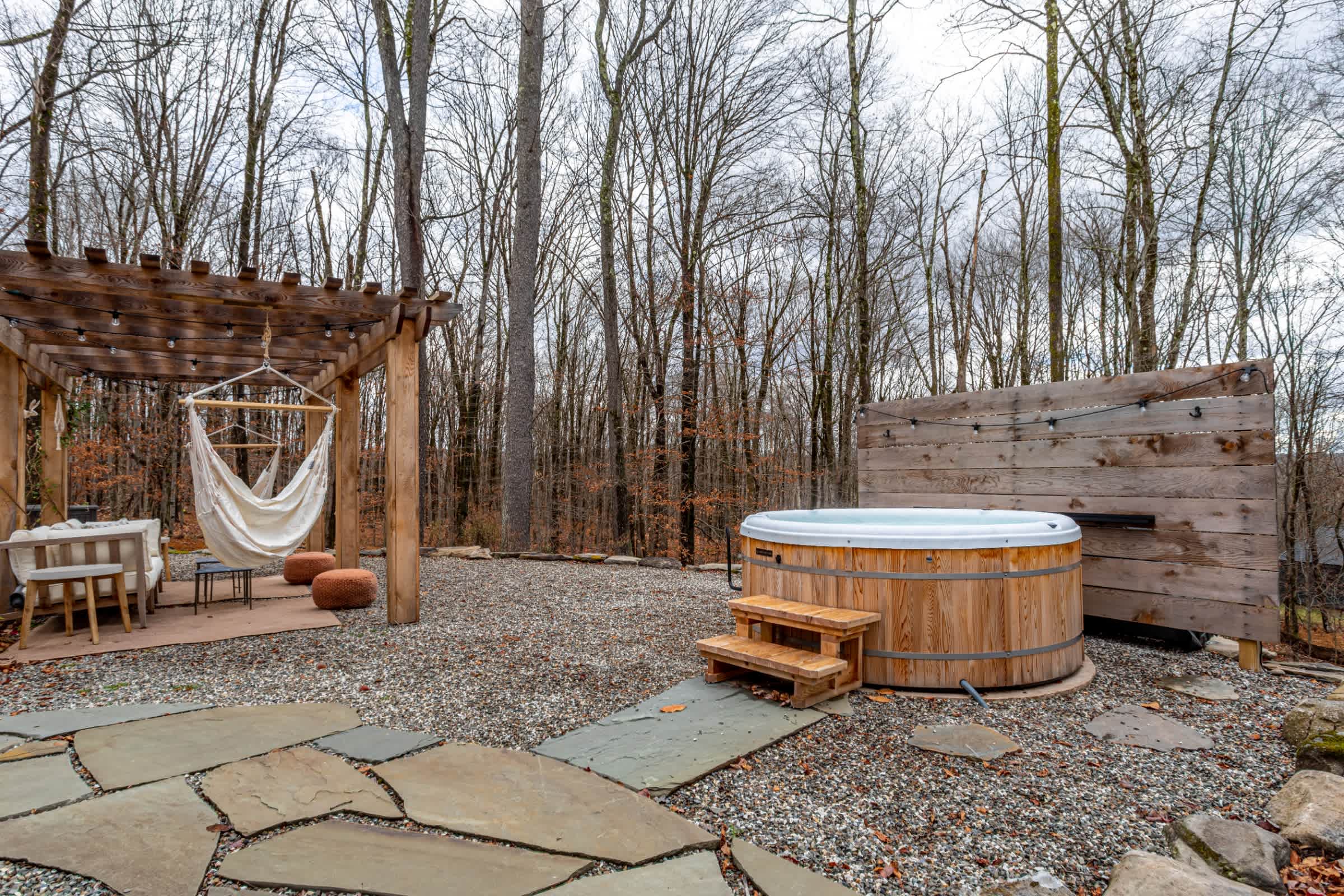 An outdoor hot tub with wooden steps is set on a gravel patio, surrounded by bare trees. A wooden privacy screen is behind the hot tub, and a pergola with a hanging hammock is visible to the left.