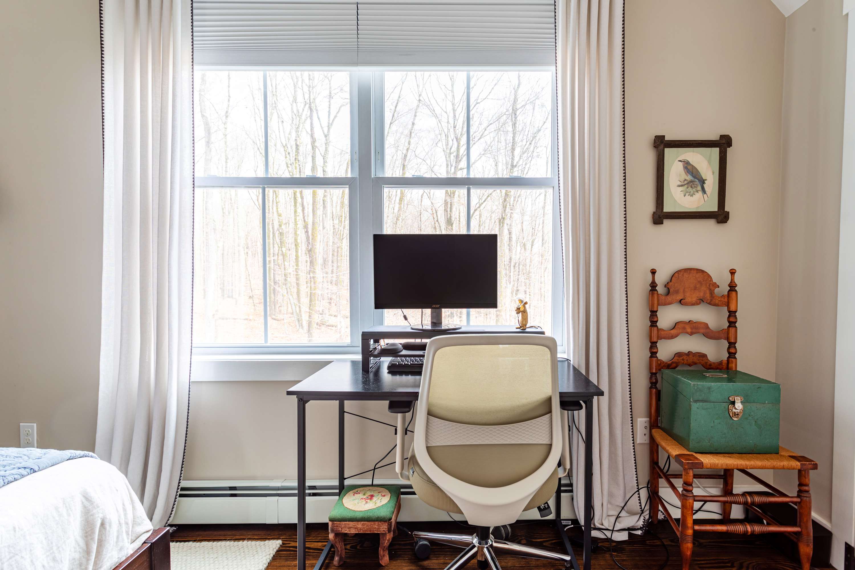 A bright bedroom features a modern desk and ergonomic chair positioned in front of a large window overlooking a bare winter forest. White curtains frame the window, and a traditional wooden chair with a green box sits beside the desk.