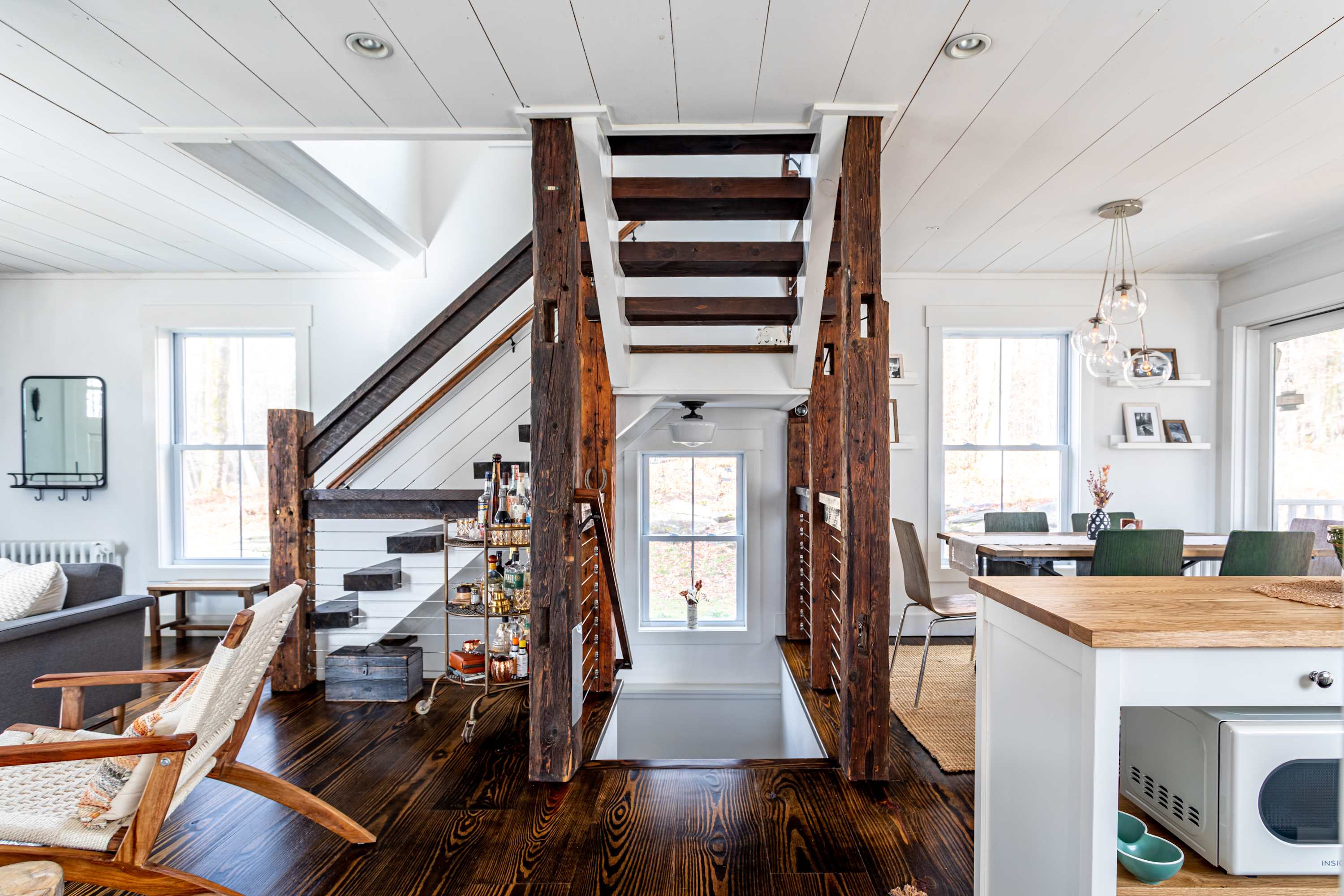 Open-concept living space showcasing a rustic wooden staircase with cable railings, dark wood floors, and a bar cart, leading into the dining and kitchen areas.