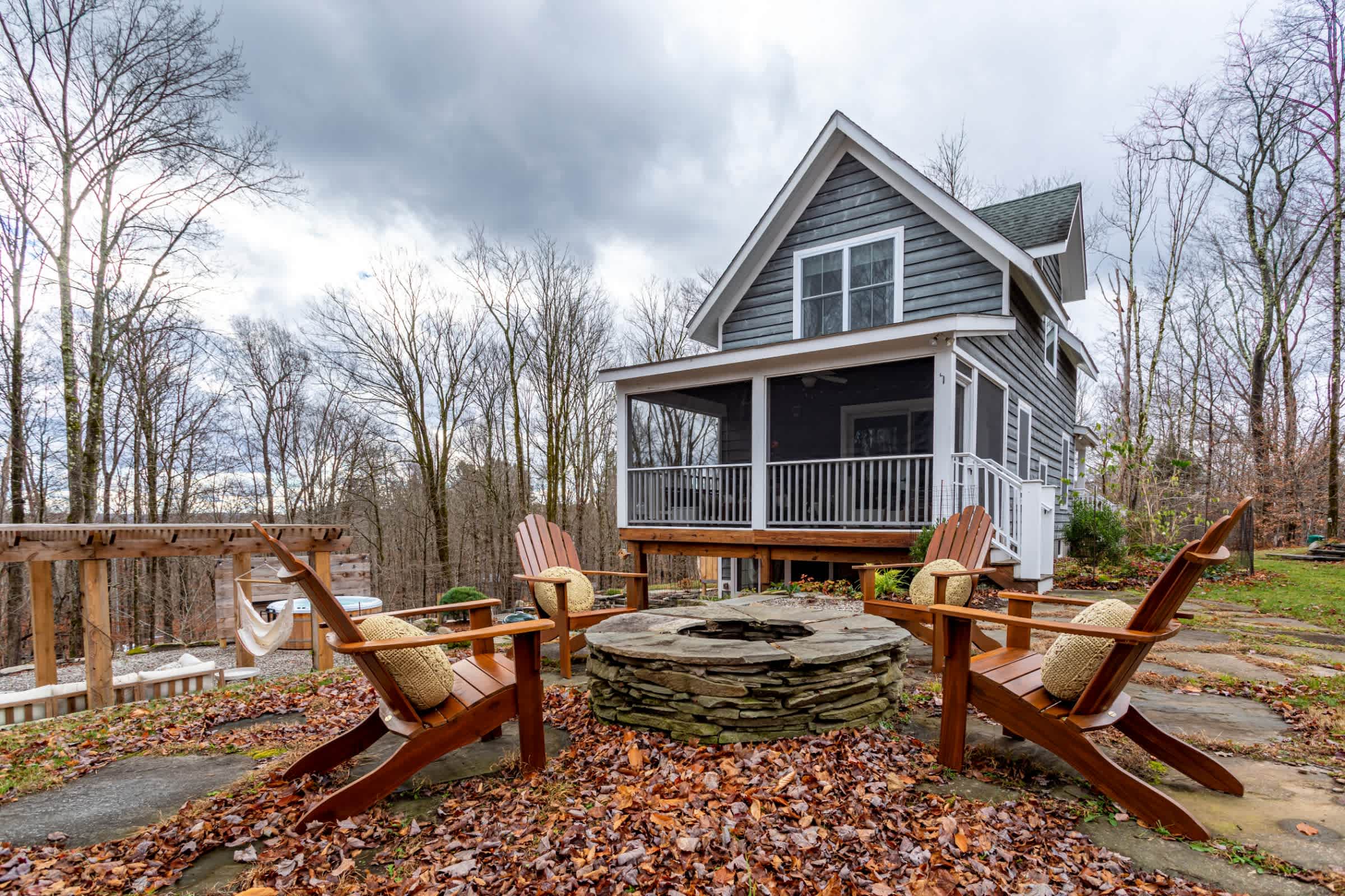 The back of a gray-sided cottage-style house with a screened porch is visible, overlooking a flagstone patio with a stone fire pit and wooden Adirondack chairs, surrounded by bare trees and fallen leaves.