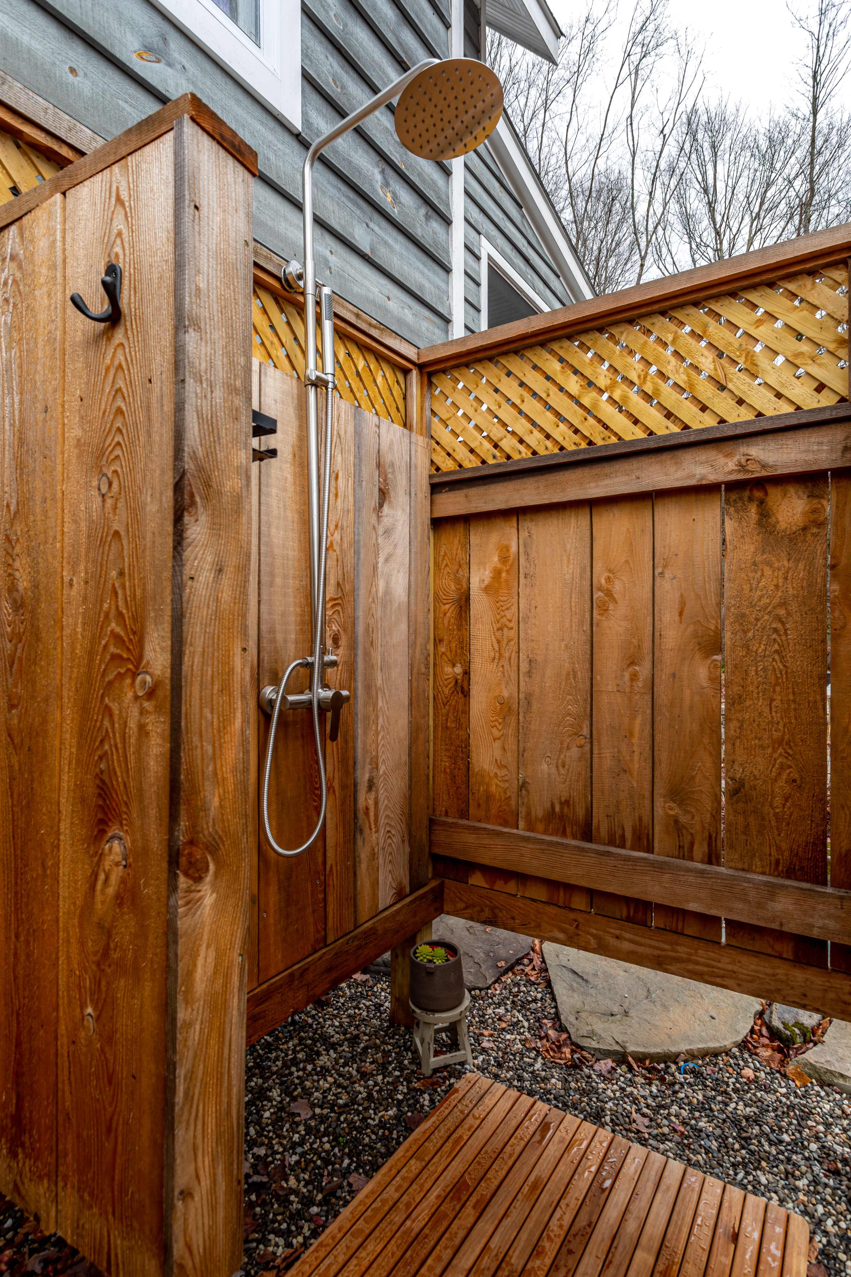 A rustic outdoor shower with wooden walls and a slatted wooden floor is built against the side of a gray-sided house. A rain shower head and handheld sprayer are visible.