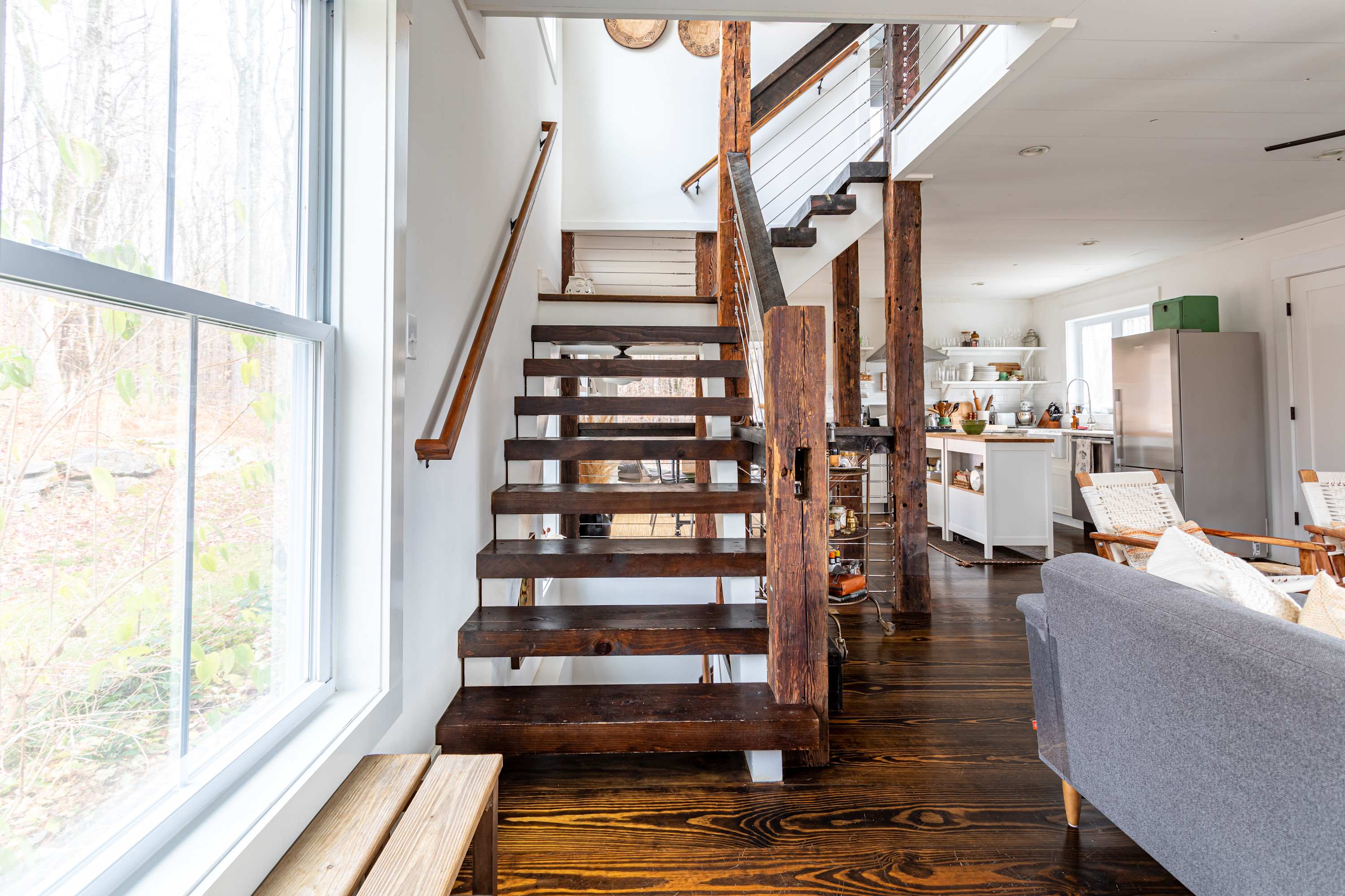 A striking open-tread wooden staircase with rustic wood posts and cable railings leads upstairs, set against white walls and dark hardwood floors, with a glimpse of an open-concept living area and kitchen beyond.