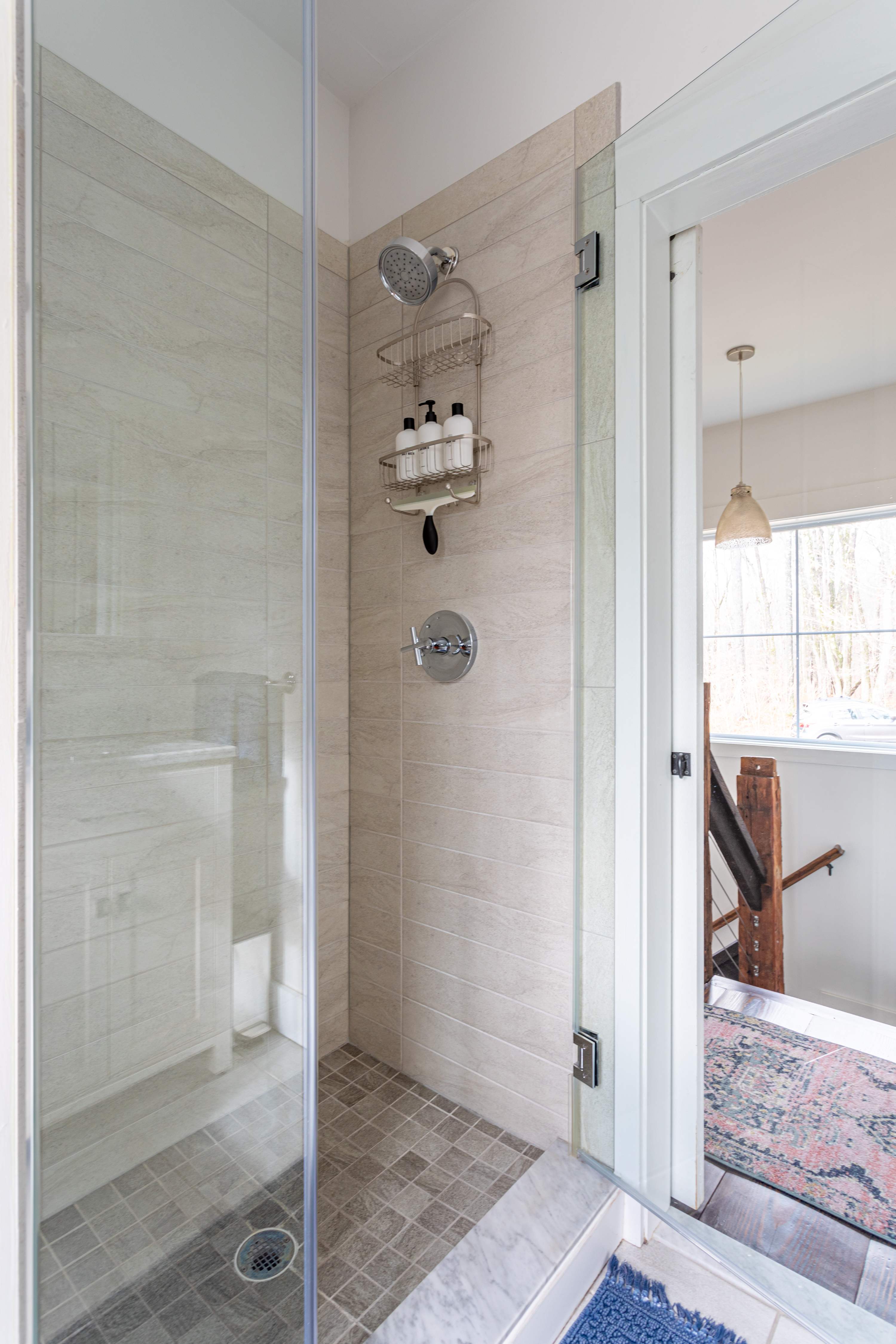 A modern walk-in shower with light-colored tiled walls and a glass enclosure, featuring a rainfall showerhead and a separate handheld sprayer, with a glimpse of the hallway beyond.