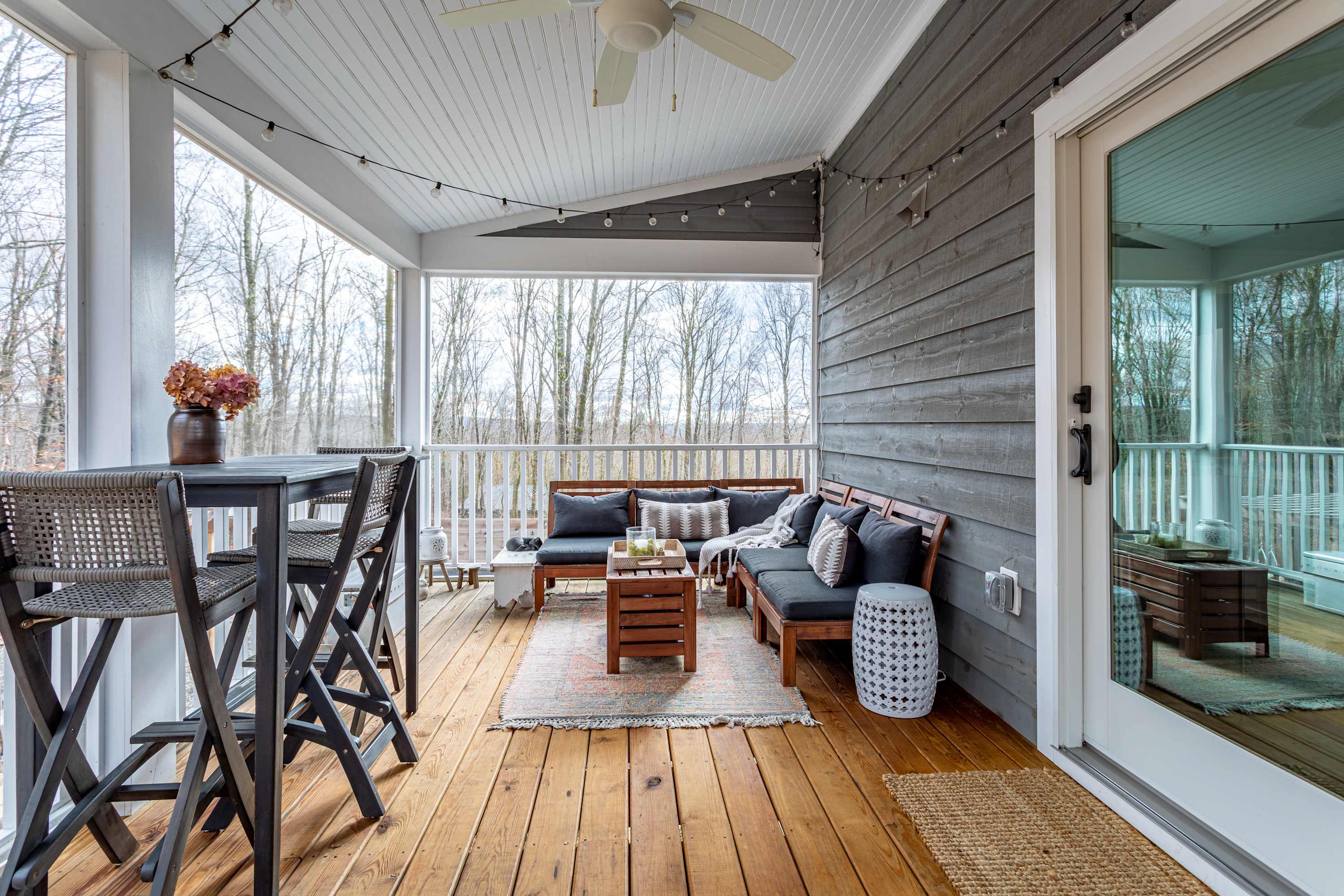 A spacious covered porch with wooden floors and gray shiplap walls, furnished with a wooden slatted sofa, a high-top dining table with stools, and string lights, offering a view of the surrounding woods.