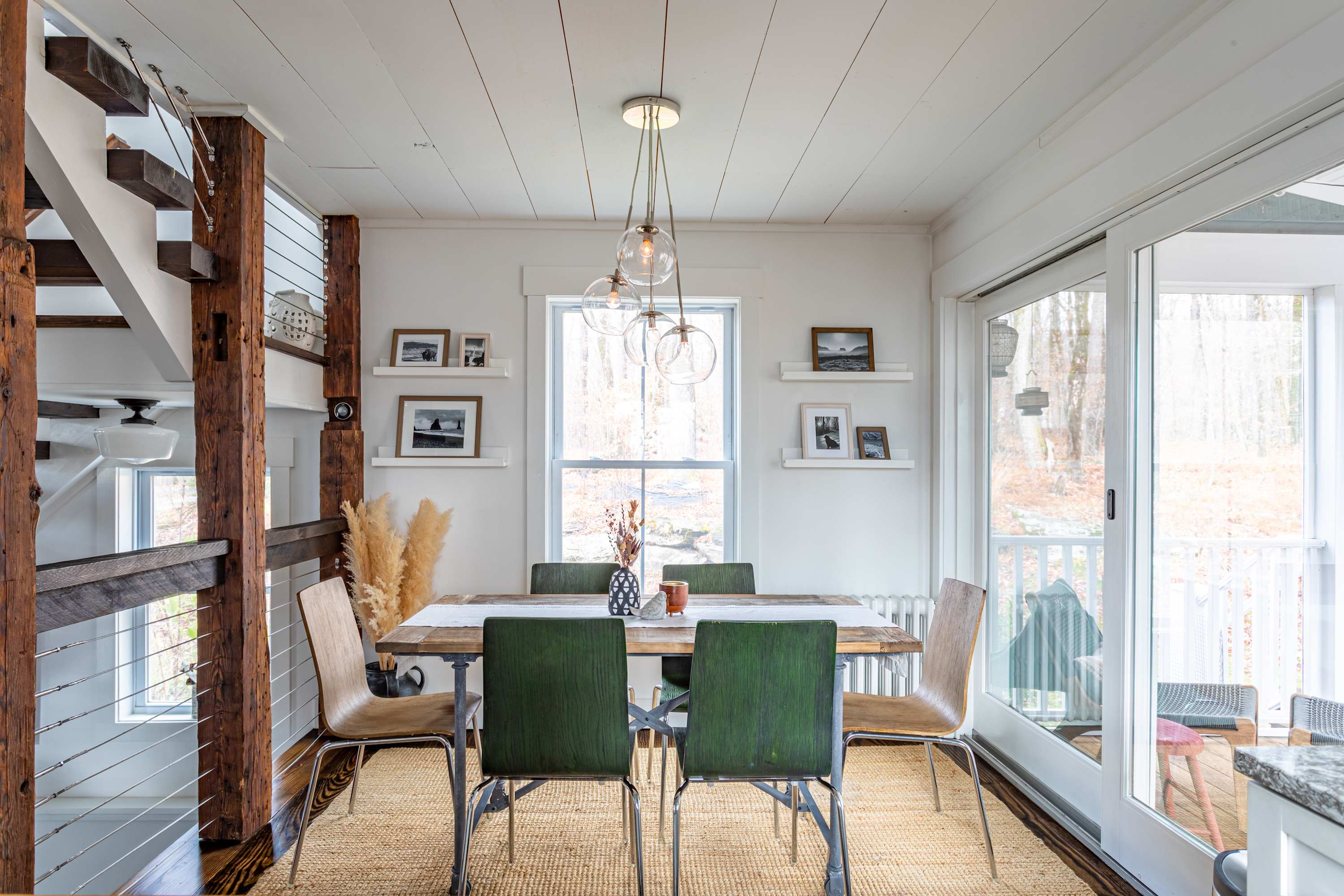 Inviting dining space with a rustic wooden table, green and wooden chairs, a modern chandelier, and decorative wall shelves, situated next to a striking wooden staircase.