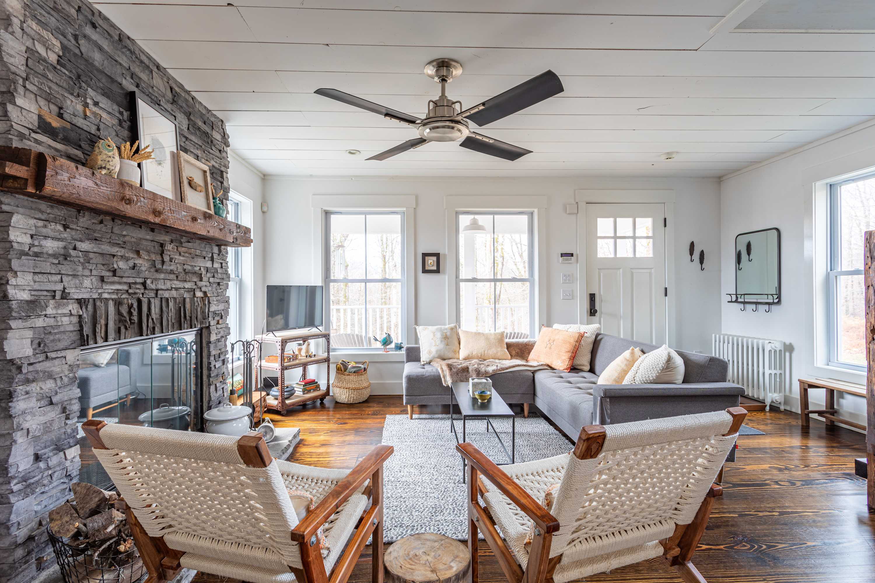 A bright living room showcases a floor-to-ceiling stone fireplace with a rustic wooden mantel, dark hardwood floors, and a white shiplap ceiling with a ceiling fan. A gray sectional sofa with throw pillows and a black coffee table define the seating area, complemented by two woven chairs.