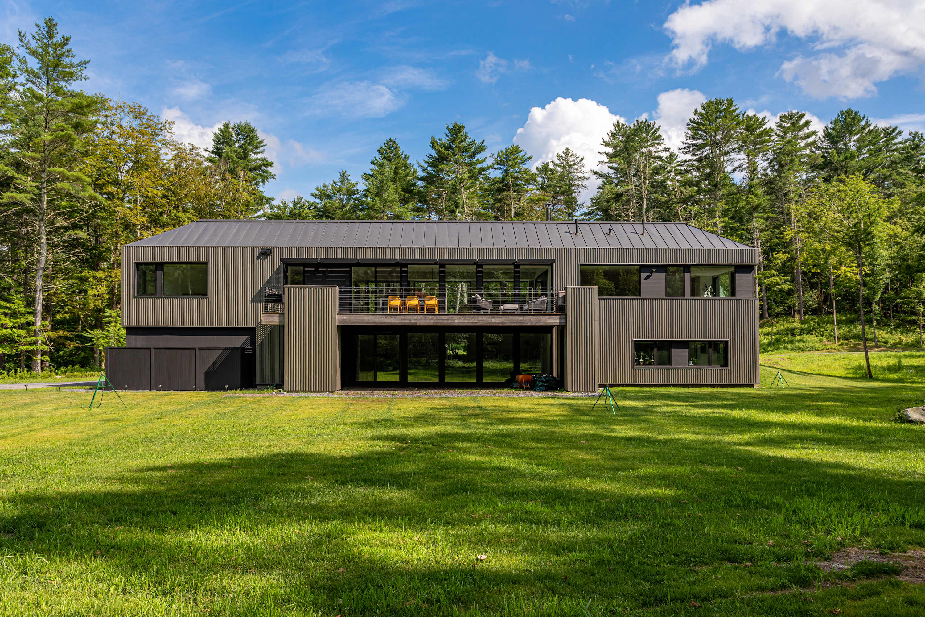 Modern two-story house with dark corrugated metal siding and extensive glass windows, featuring a large balcony on the upper level overlooking a spacious green lawn and dense forest.