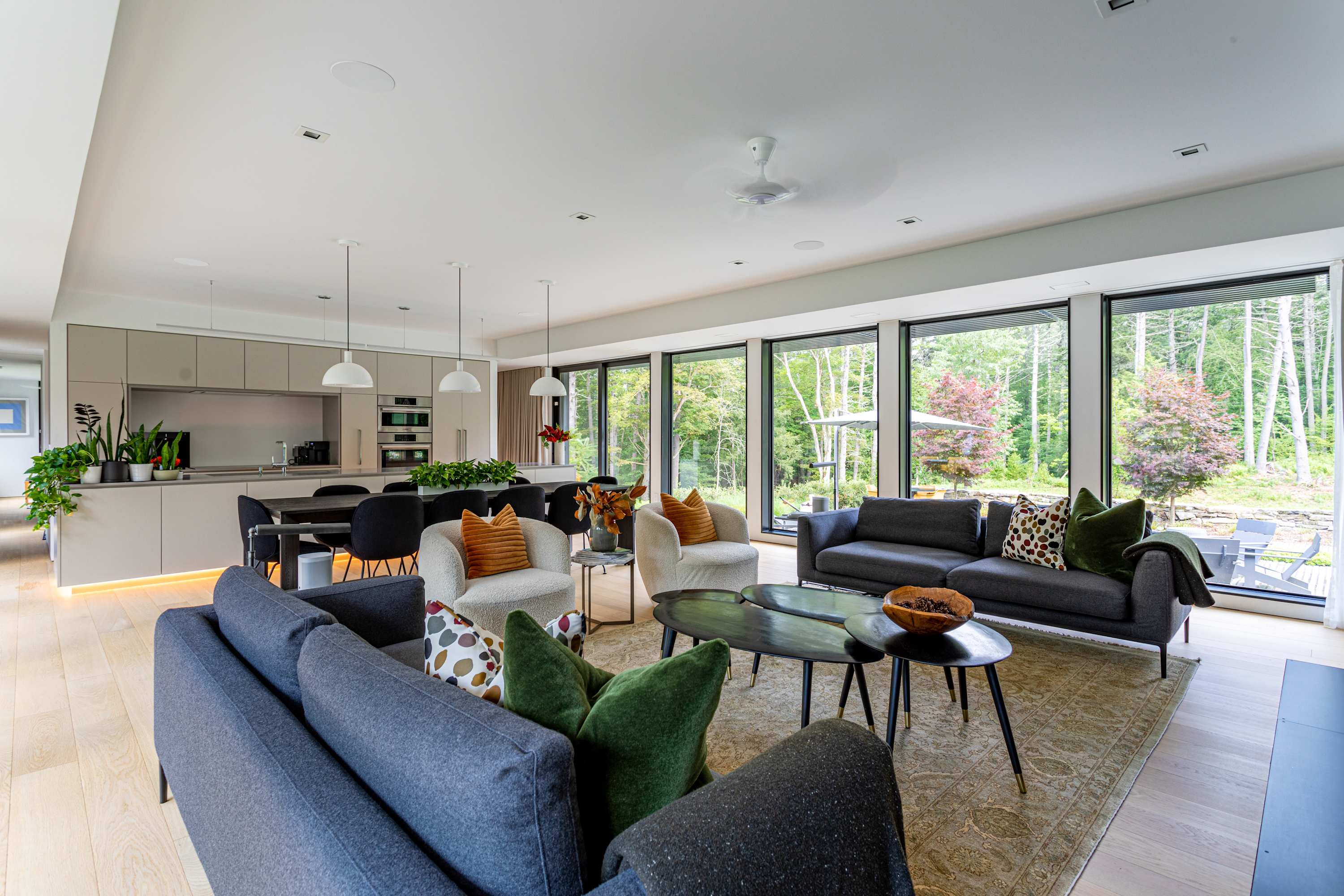 Wide view of an open-concept living area with light hardwood floors, showing two gray sofas and two white armchairs, a dining table, and a modern kitchen, all bathed in natural light from expansive windows facing the forest.