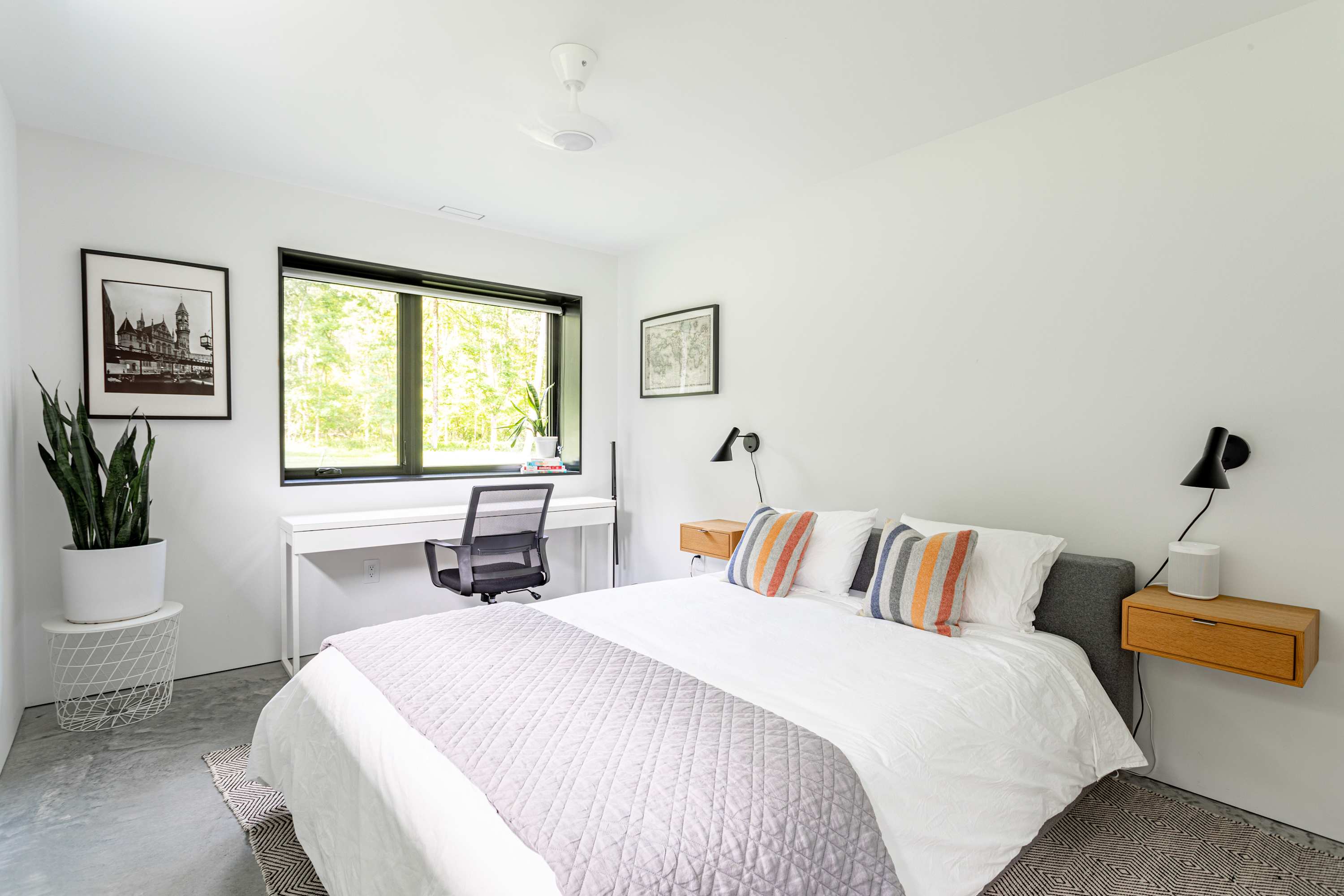 Modern bedroom with a white queen-sized bed, a grey quilted throw, and colorful striped pillows. A white desk and office chair are positioned by a large window overlooking greenery, with a snake plant in a white pot nearby.