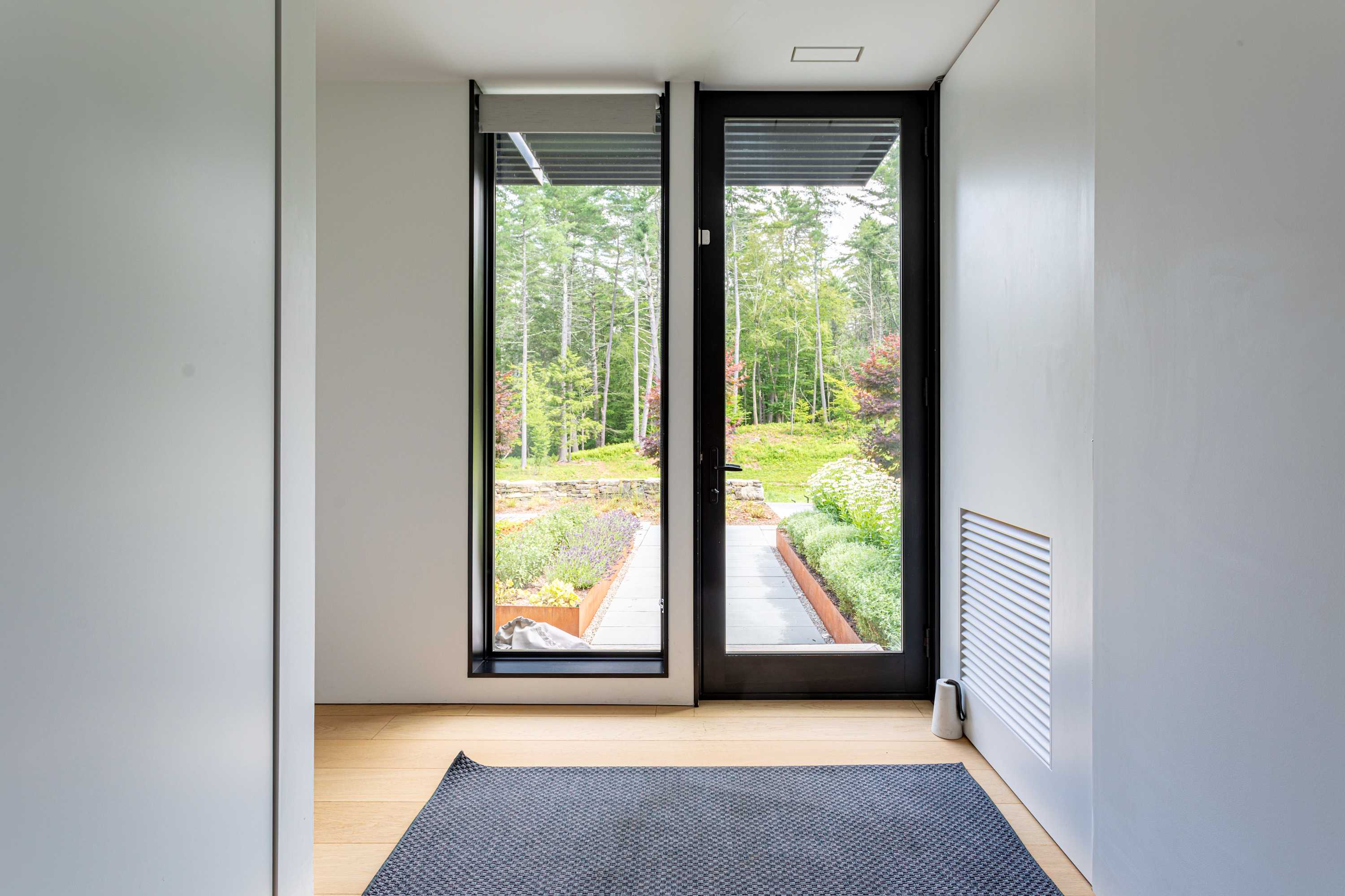 A bright hallway with light hardwood floors and a dark patterned rug, leading to a glass-paned exterior door and large window overlooking a landscaped garden. White walls and minimalist design characterize the space.