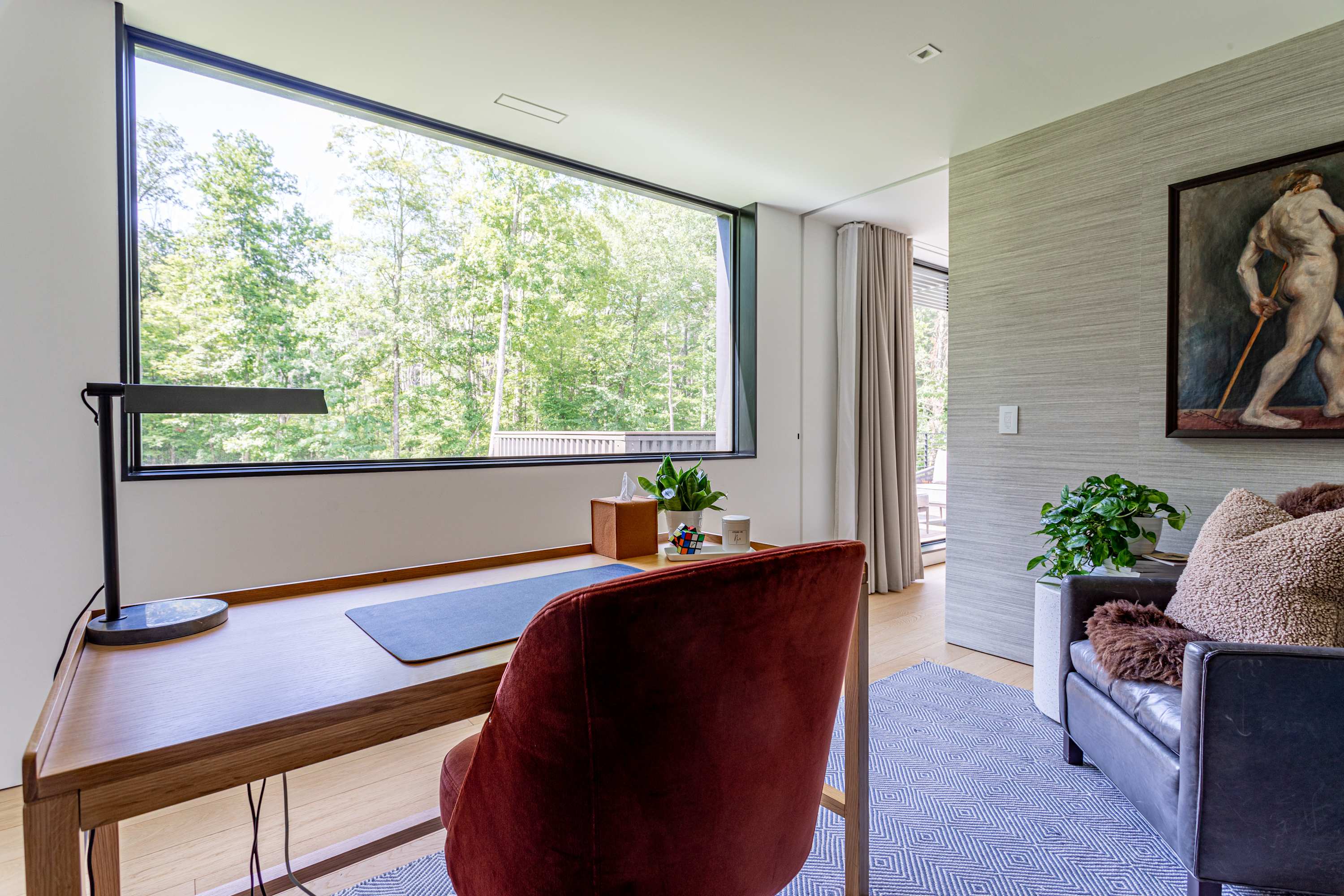 The home office from another angle, showcasing a large horizontal window offering expansive views of lush green trees. A wooden desk with a black task lamp and a red velvet chair are prominent, with a dark leather armchair in the corner.