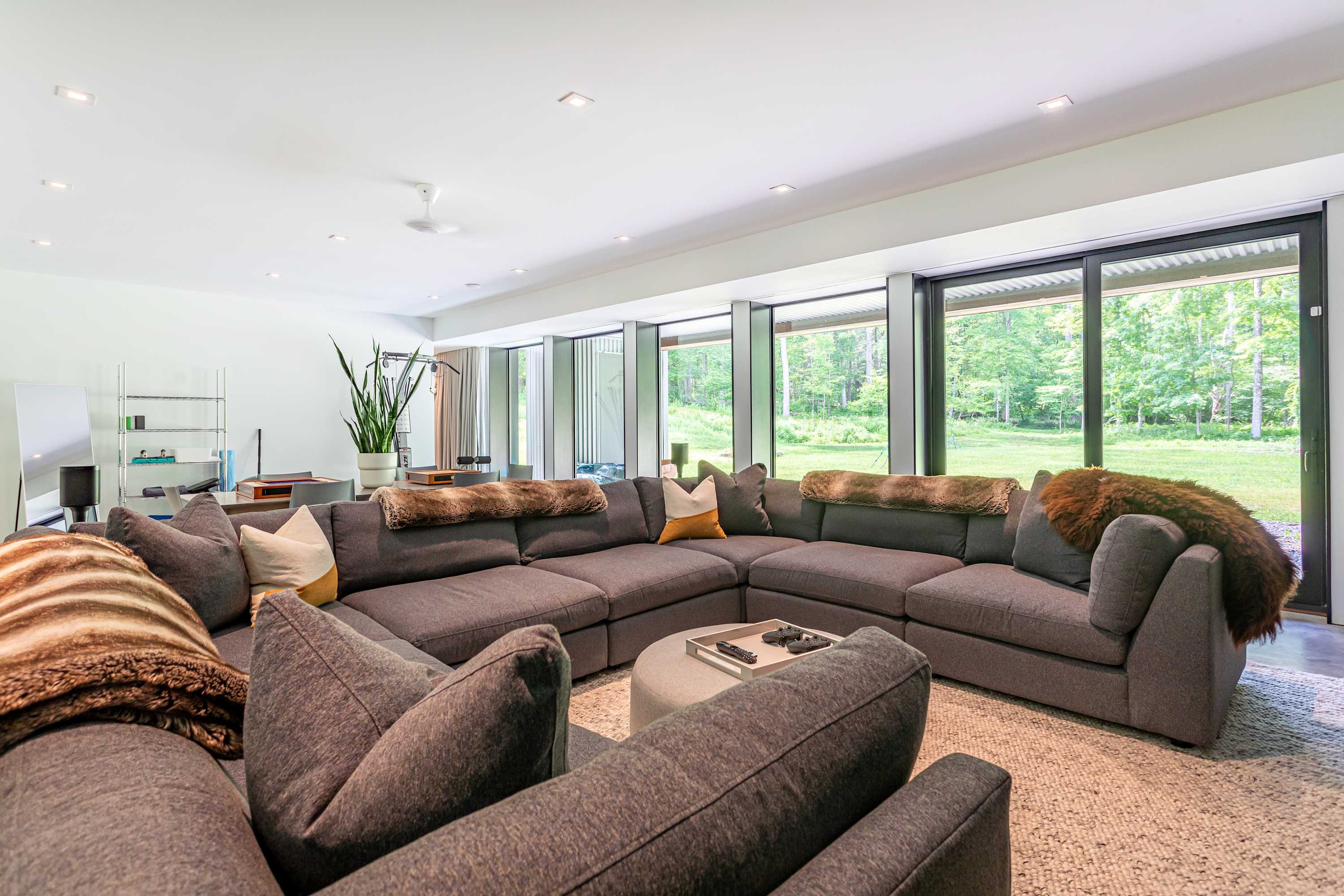 Spacious living room featuring a large grey sectional sofa adorned with faux fur throws and accent pillows. Floor-to-ceiling windows offer a panoramic view of the outdoors, and a round ottoman serves as a coffee table.
