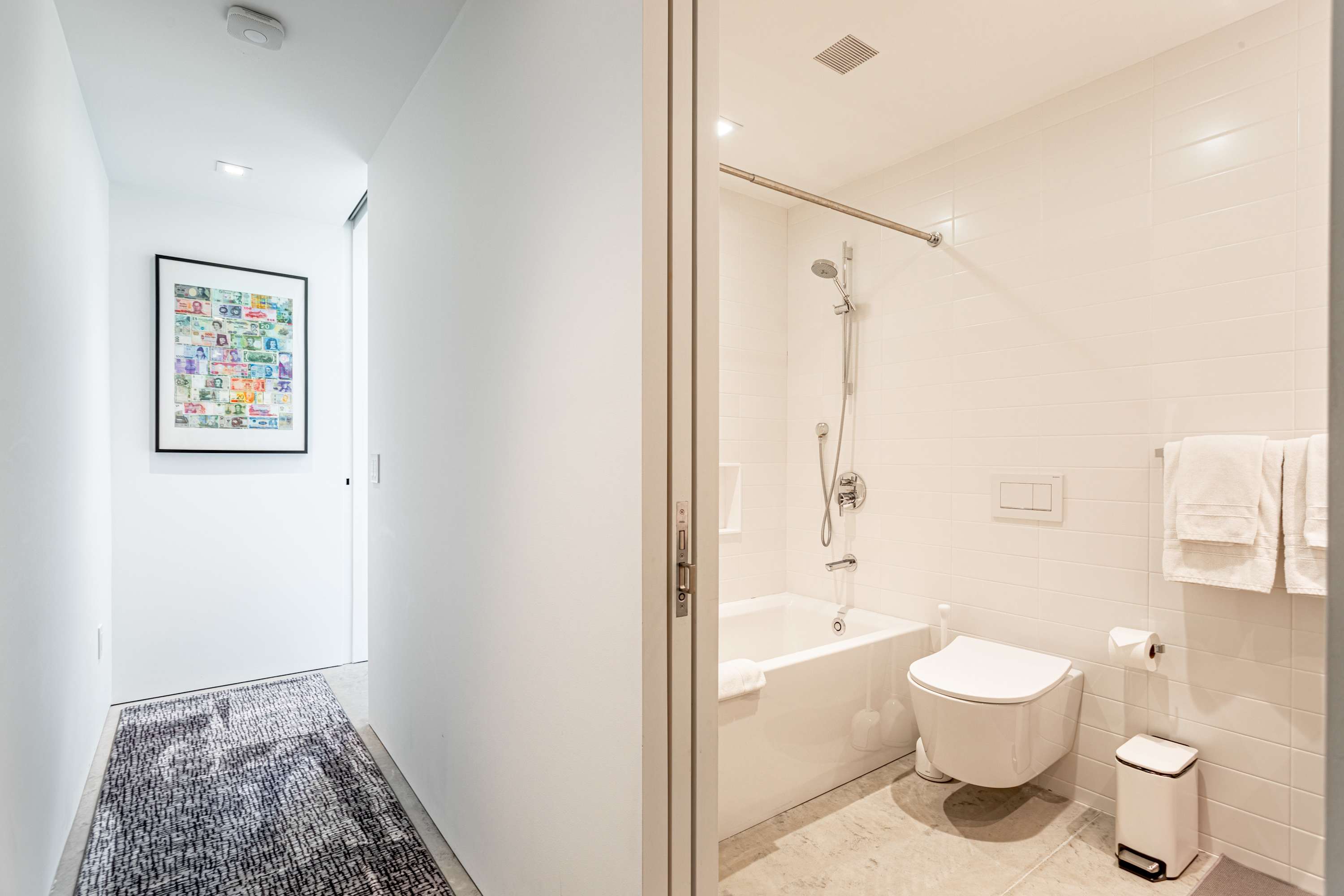 A bright, modern bathroom with white subway tile walls, a white toilet, and a shower-over-tub setup. A small white trash can and folded white towels are visible, with a hallway leading to another room.