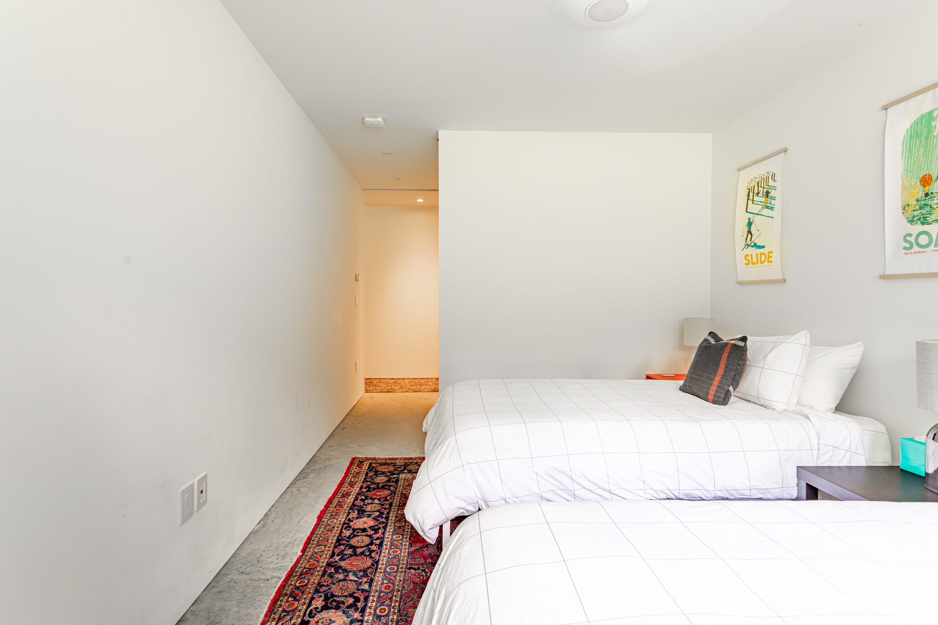 A minimalist bedroom with two queen beds featuring white grid-patterned duvets, concrete floors, and a colorful Persian rug. Two framed art pieces hang on the white wall, and a narrow hallway leads to another room.