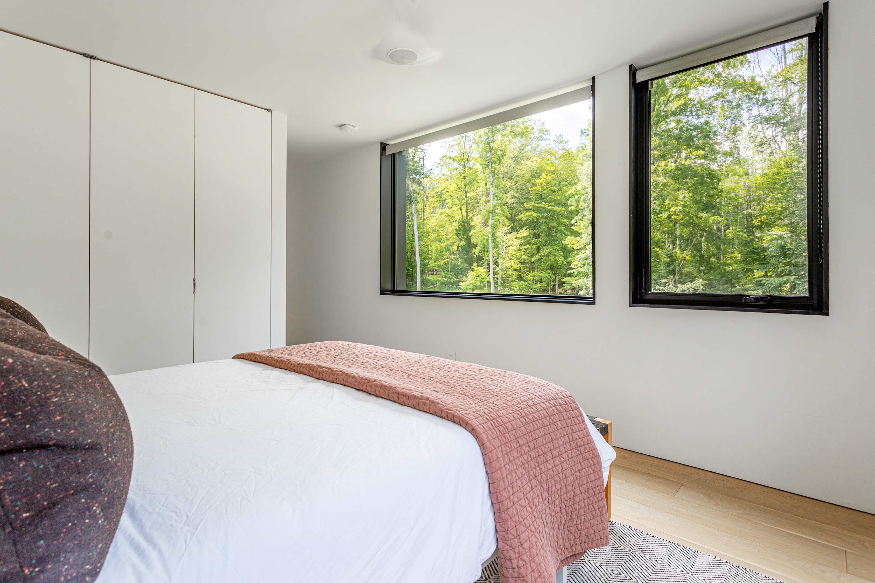 View of the contemporary bedroom showcasing two large windows with black frames, offering expansive views of the surrounding forest. White built-in wardrobes line one wall, and a queen-sized bed with a pink throw is partially visible.