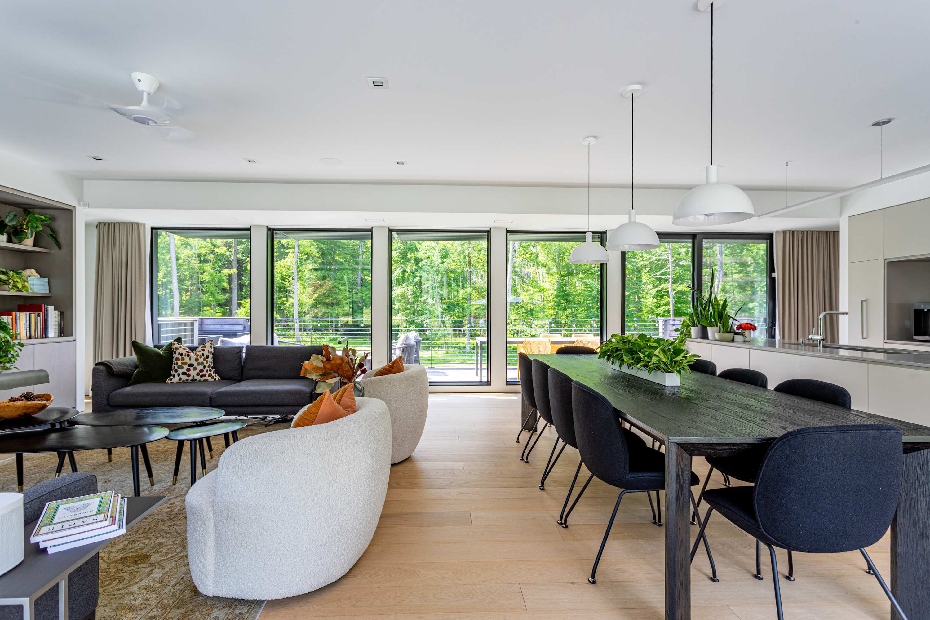 Bright open-concept living space with light hardwood floors, featuring two gray sofas and two white armchairs arranged around coffee tables, with a dining area and modern kitchen visible in the background, all overlooking a forest through large windows.