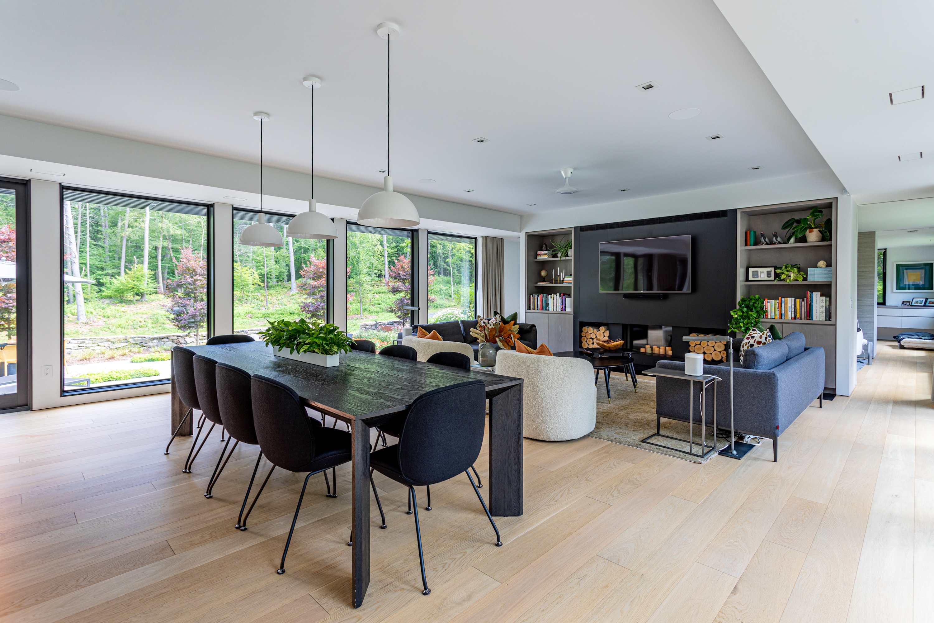An expansive open-plan room with a dark wooden dining table and black chairs in the foreground, leading to a living area with a dark feature wall, fireplace, and built-in shelving. Floor-to-ceiling windows line the wall, showcasing a vibrant green outdoor landscape.
