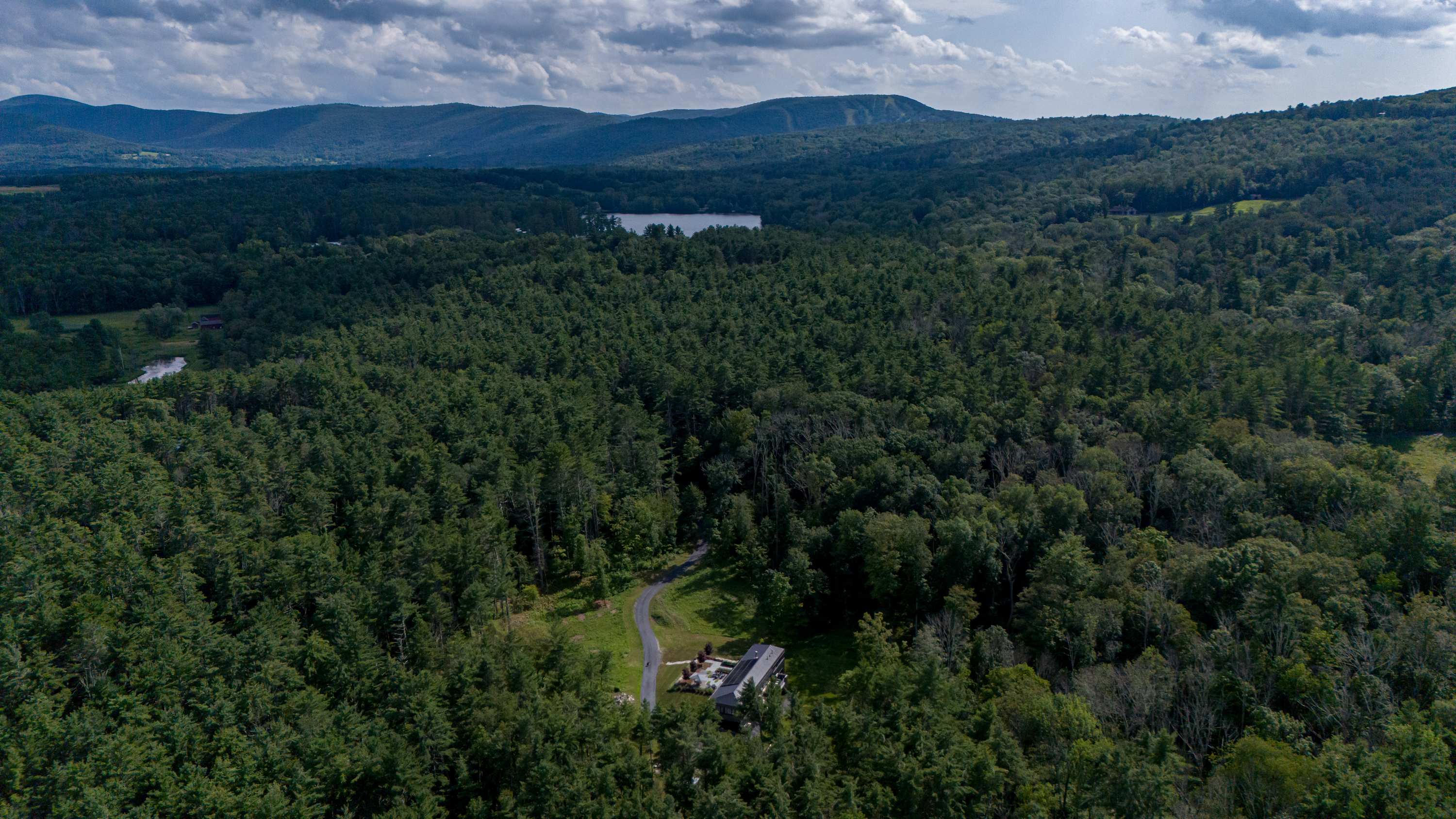 An expansive aerial view captures the modern house and its winding gravel driveway, enveloped by vast, dense green forests, with distant mountains and a serene lake visible under a partly cloudy sky.