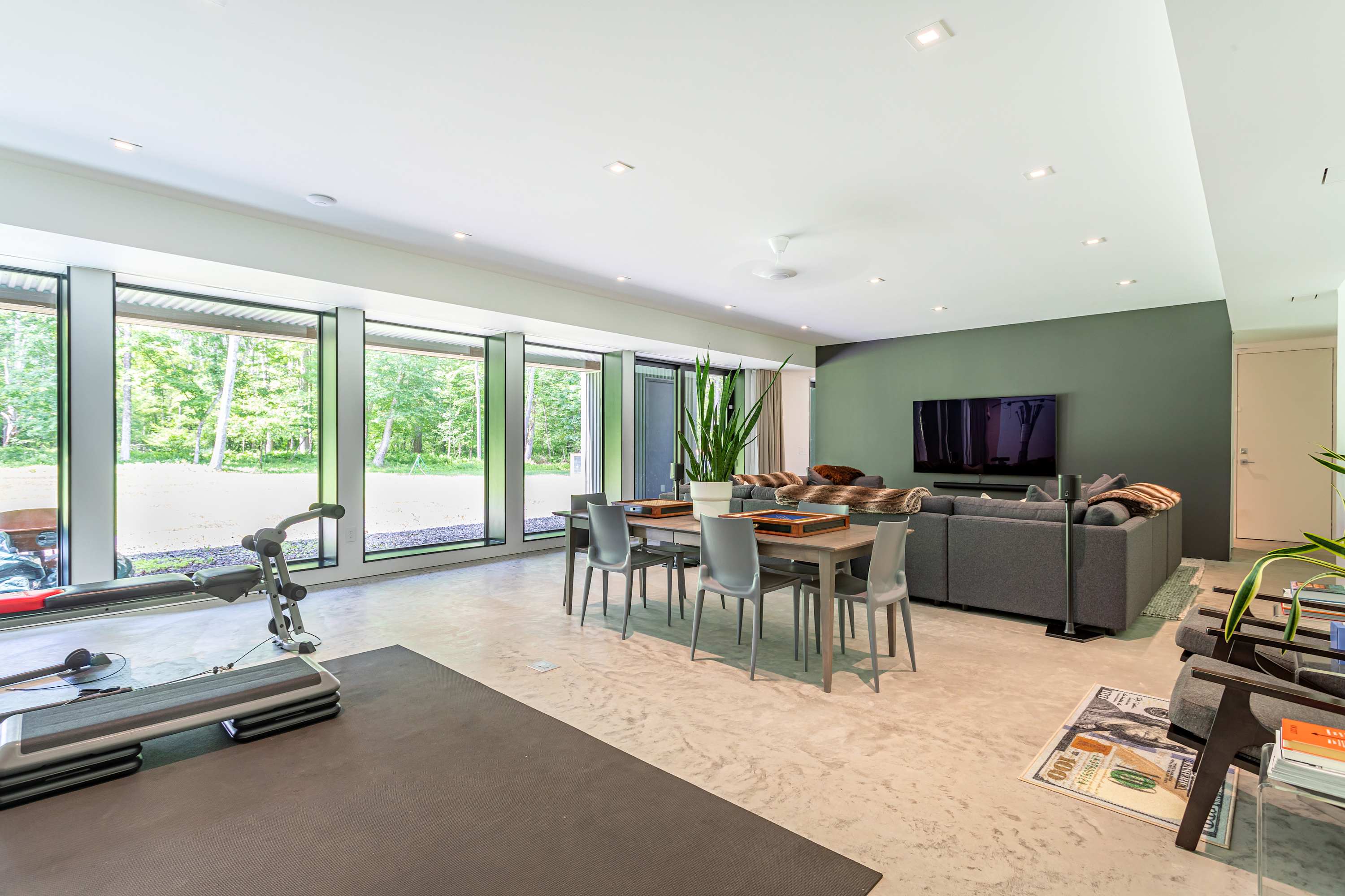 Expansive open-plan living area combining a comfortable grey sectional sofa facing a wall-mounted TV, a dining/game table, and a home gym setup. Floor-to-ceiling windows span the length of the room, offering views of the surrounding nature.