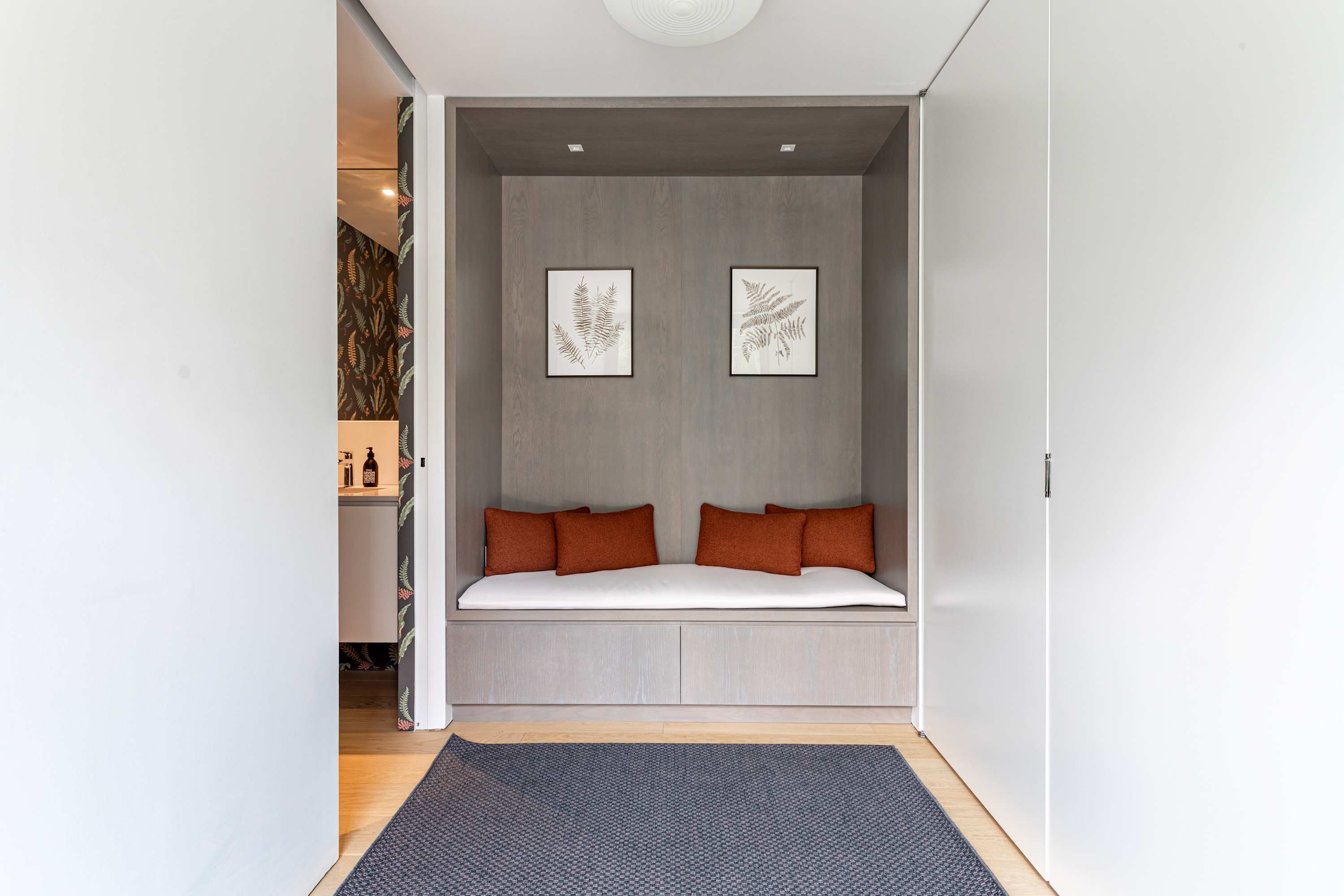 A cozy built-in nook in a hallway, featuring a light wood bench with a white cushion and orange throw pillows, flanked by two framed botanical prints. The space has light hardwood floors and white walls.