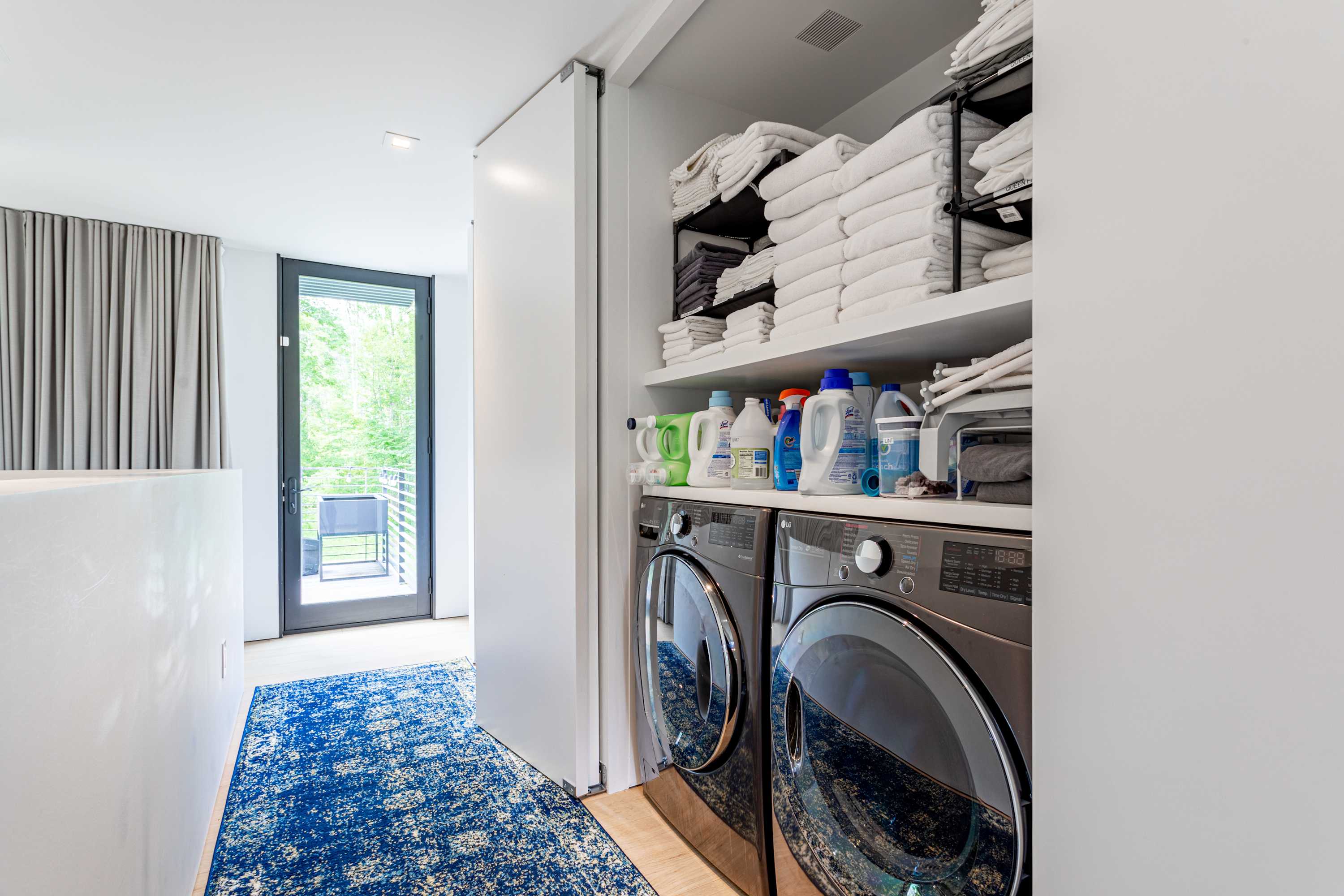A dedicated laundry area with stacked front-loading washer and dryer units, neatly organized shelves filled with white towels and cleaning supplies. A blue patterned rug covers the hardwood floor in the adjacent hallway.