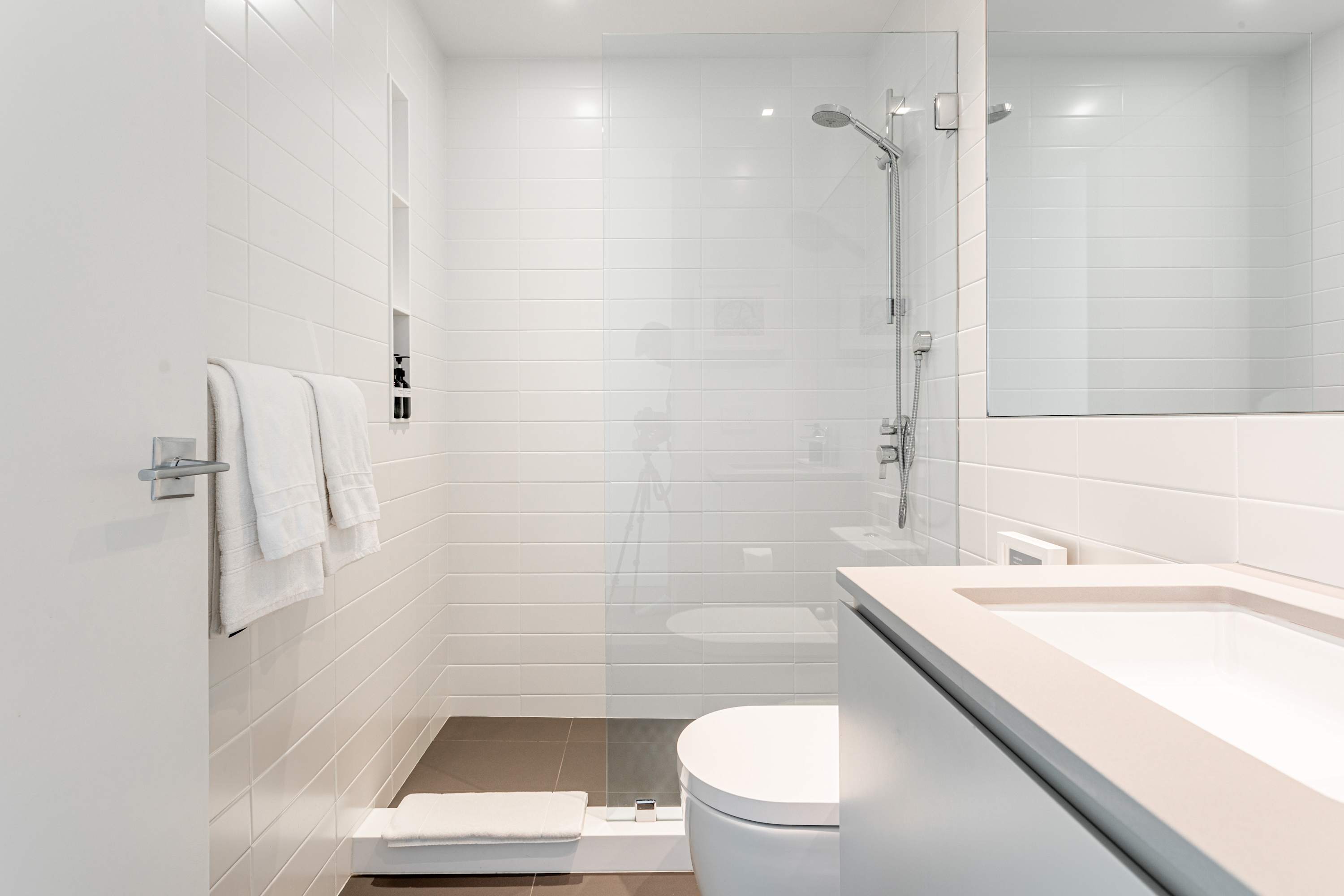 Bright, modern bathroom with white subway tile walls and a glass-enclosed shower. A white floating vanity with an integrated sink and a large mirror is on the right, and a toilet is visible next to the shower.