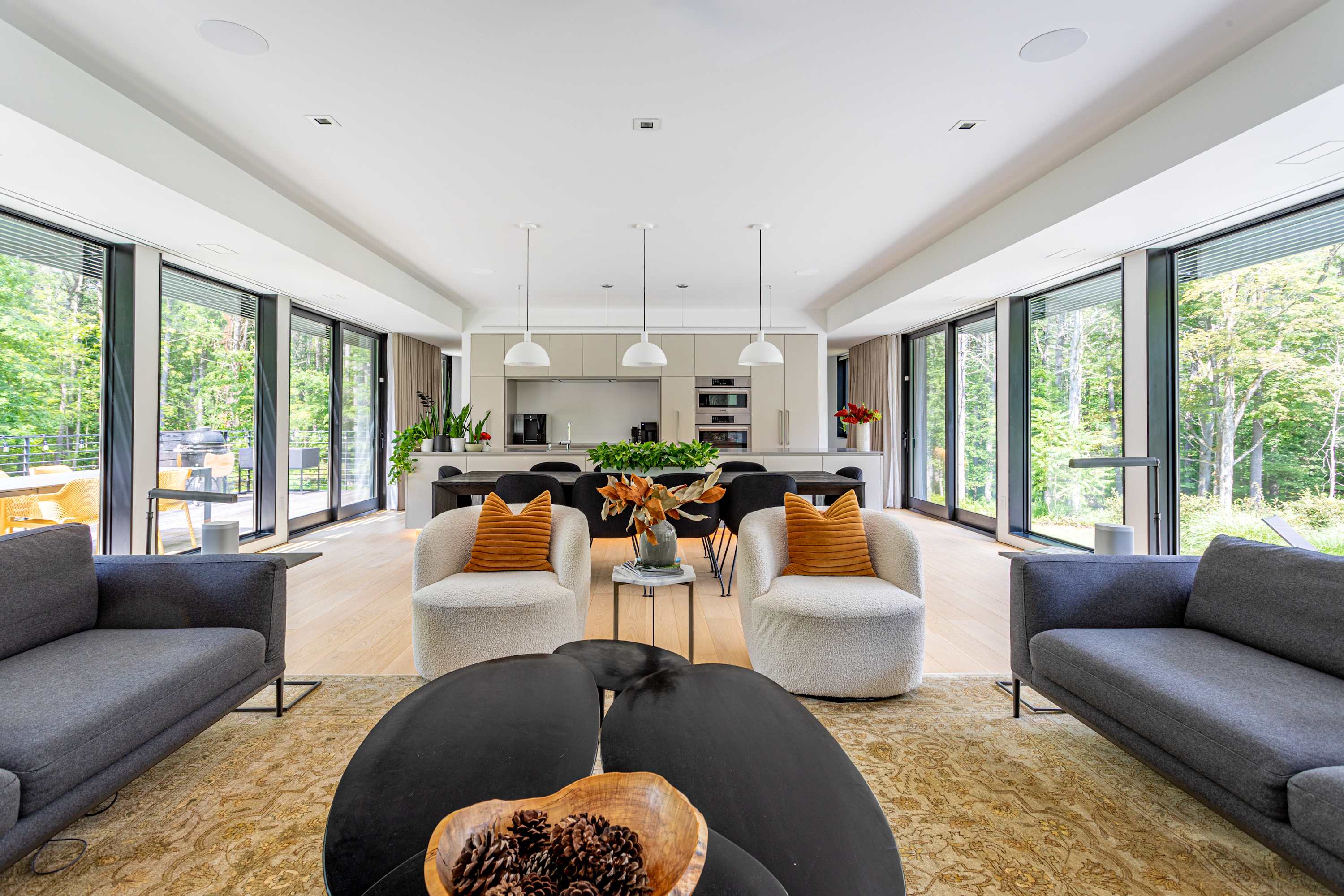A contemporary open-concept space featuring two cream-colored armchairs with orange pillows, a dark coffee table, and a patterned rug in the foreground. The background reveals a sleek kitchen with light grey cabinets and a dining table, all surrounded by floor-to-ceiling windows.