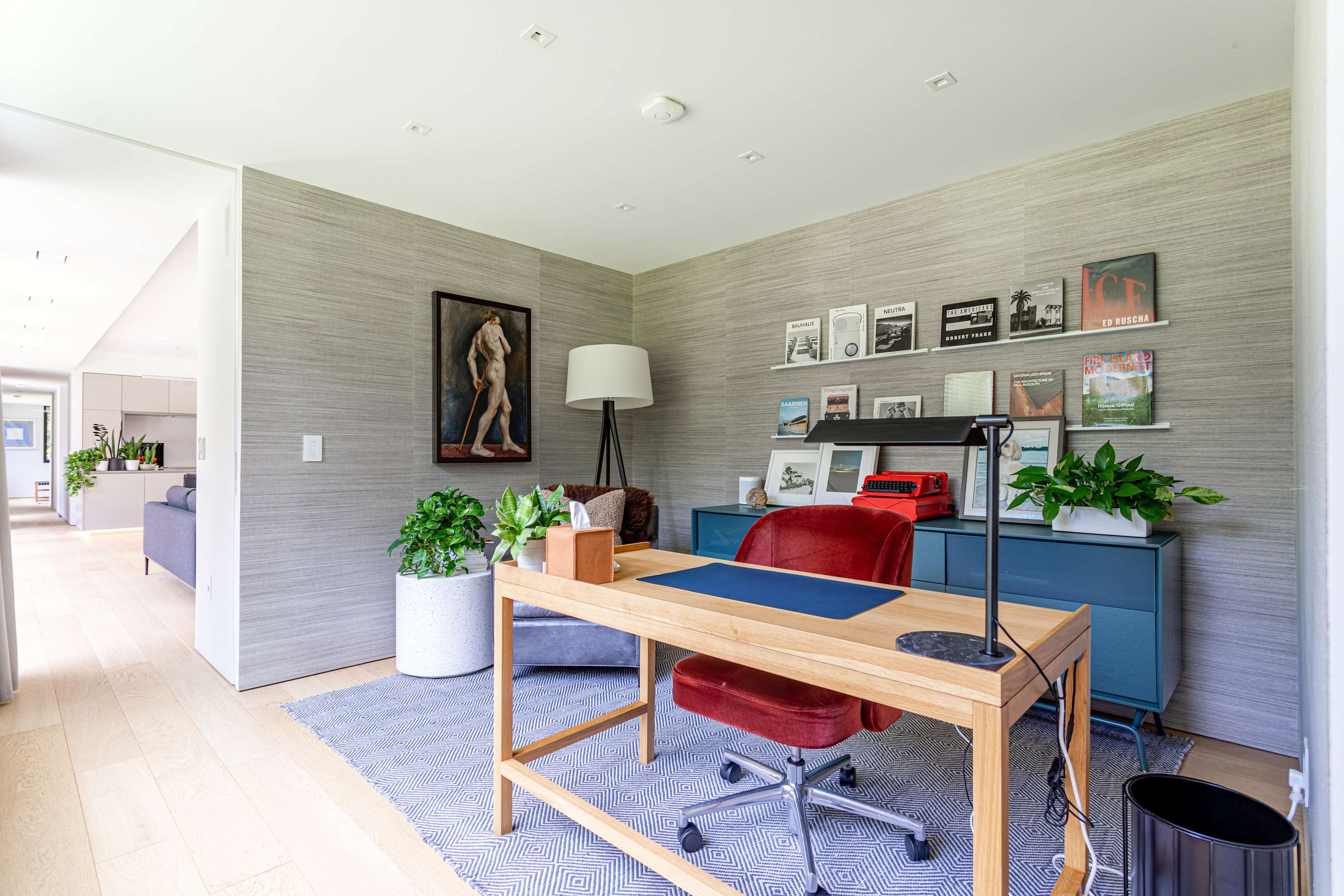 A stylish home office featuring a light wood desk with a red velvet chair, set against a textured grey wallpaper. Floating shelves display books and decor, and a large painting of a figure hangs on the wall.