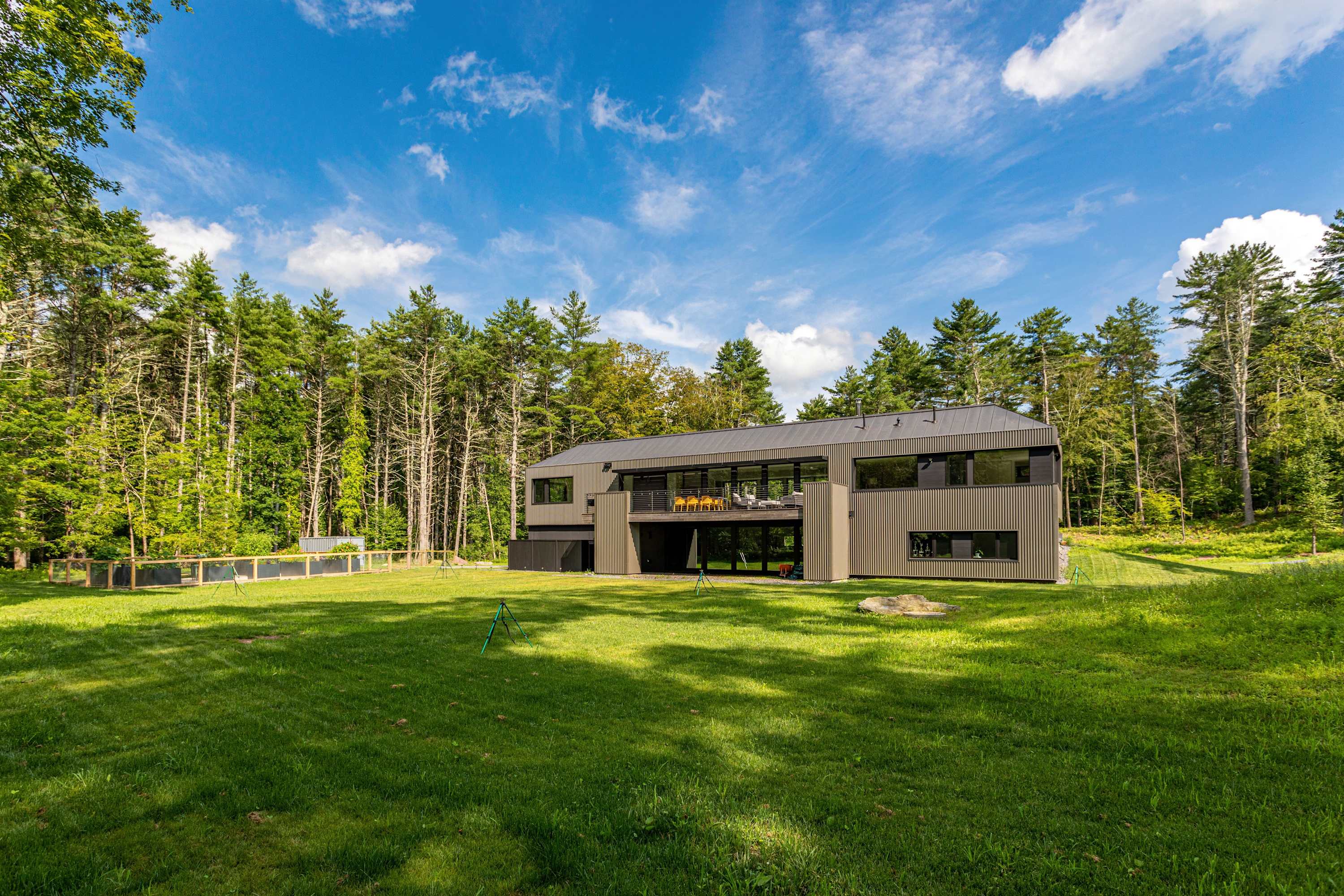 The modern back exterior of the house, featuring dark grey siding and large windows, set against a lush green lawn and dense forest. A covered balcony with seating is visible on the upper level.