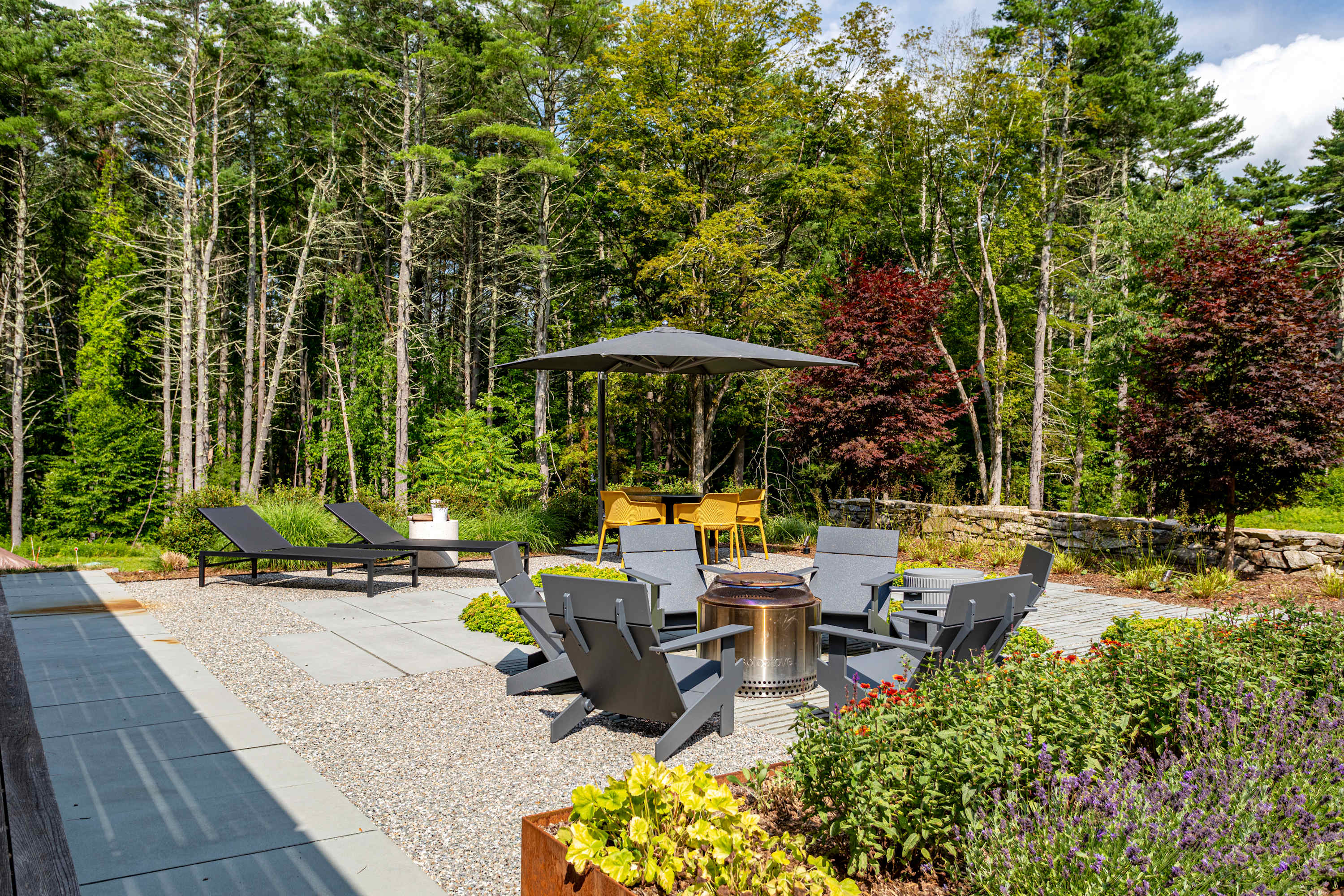 Outdoor patio area with a central fire pit surrounded by gray Adirondack chairs, set on gravel and concrete pavers, with raised garden beds and lush forest in the background, offering a cozy gathering space.