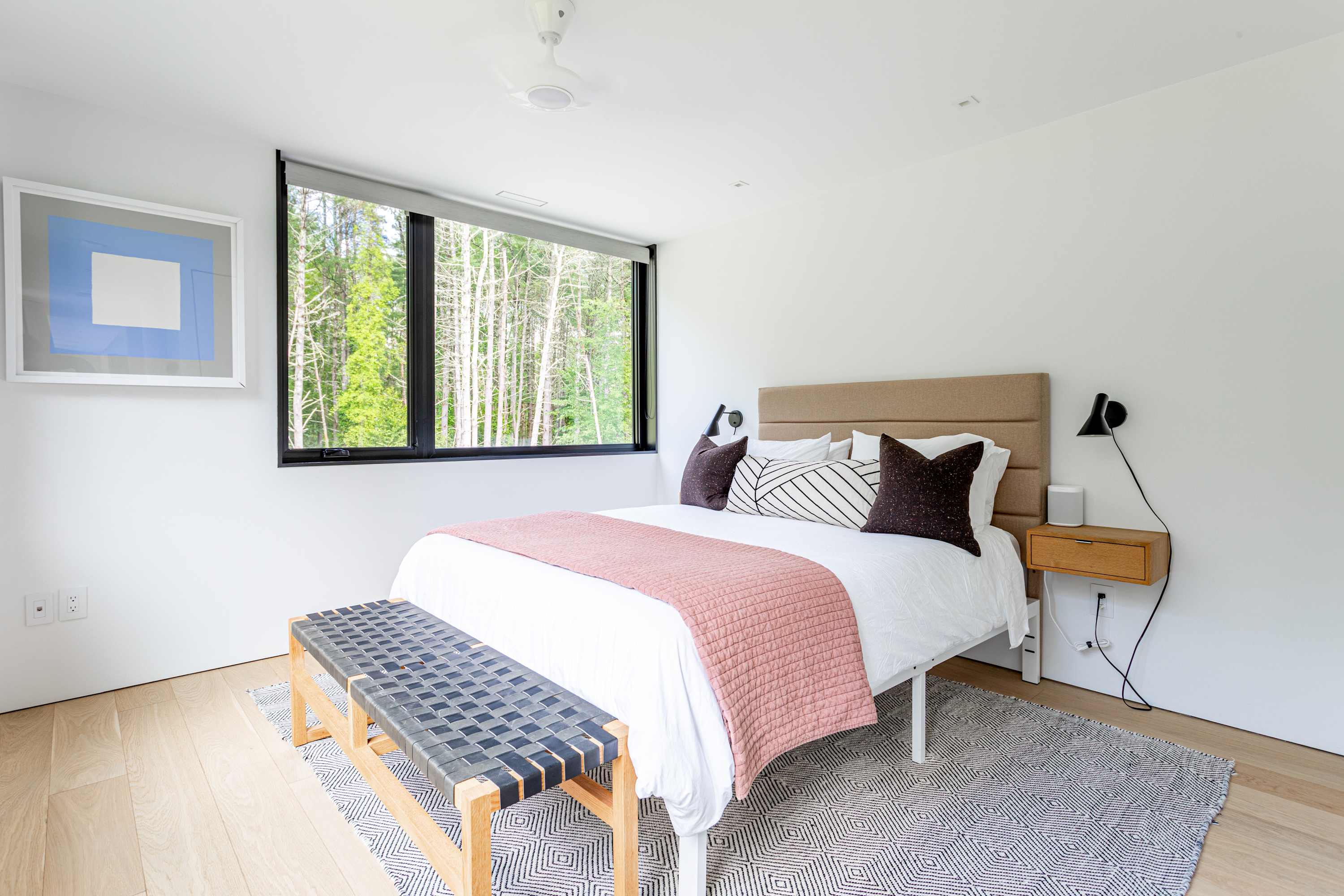 Contemporary bedroom featuring a queen-sized bed with a tan upholstered headboard, white bedding, and a pink quilted throw. A woven leather bench sits at the foot of the bed, and a large window offers views of lush trees.
