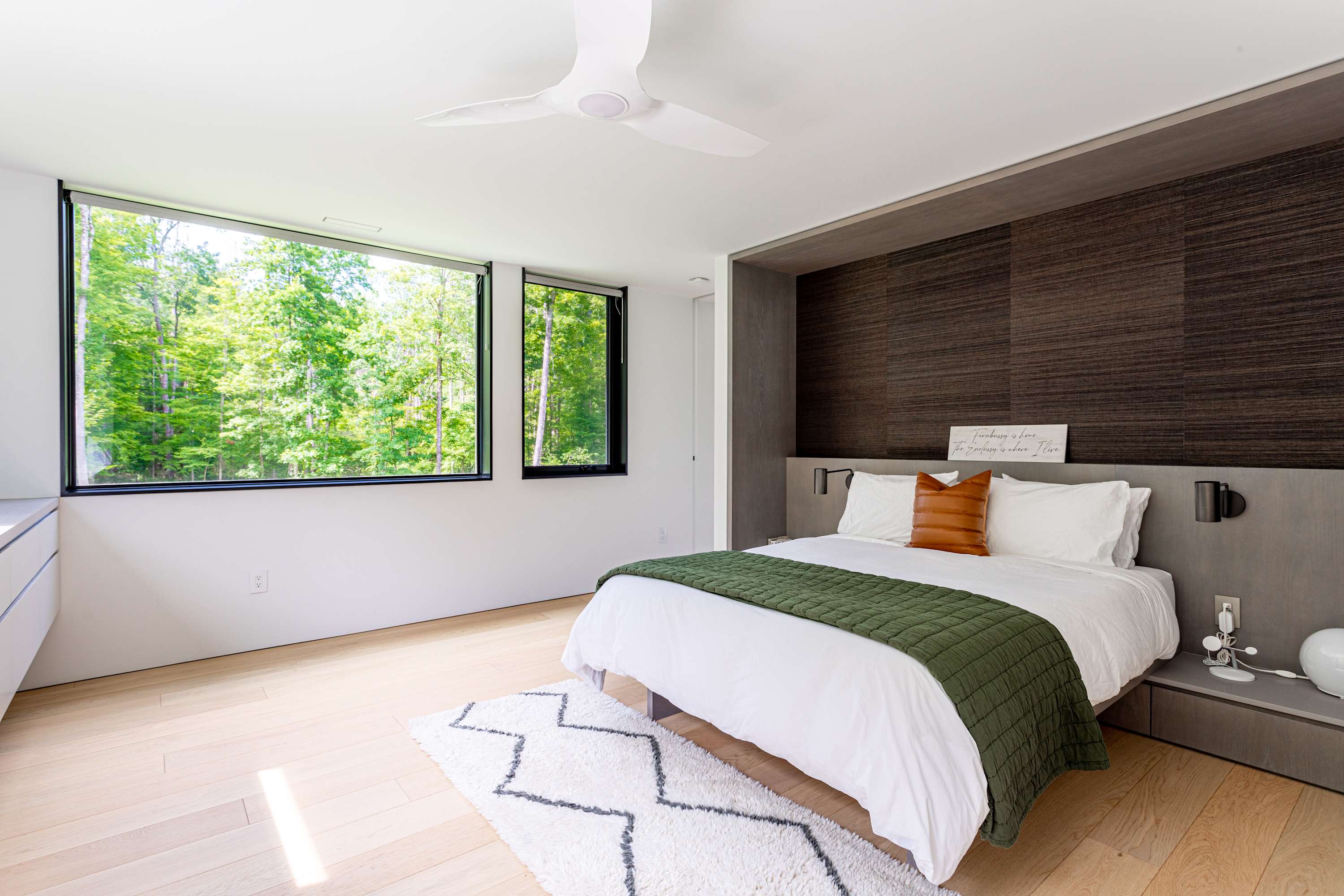 Bedroom-1 features a queen-sized bed with white bedding and a green quilted throw, set against a dark wood accent wall with integrated nightstands. Large windows provide a serene view of the surrounding forest, and light hardwood floors complete the modern aesthetic.