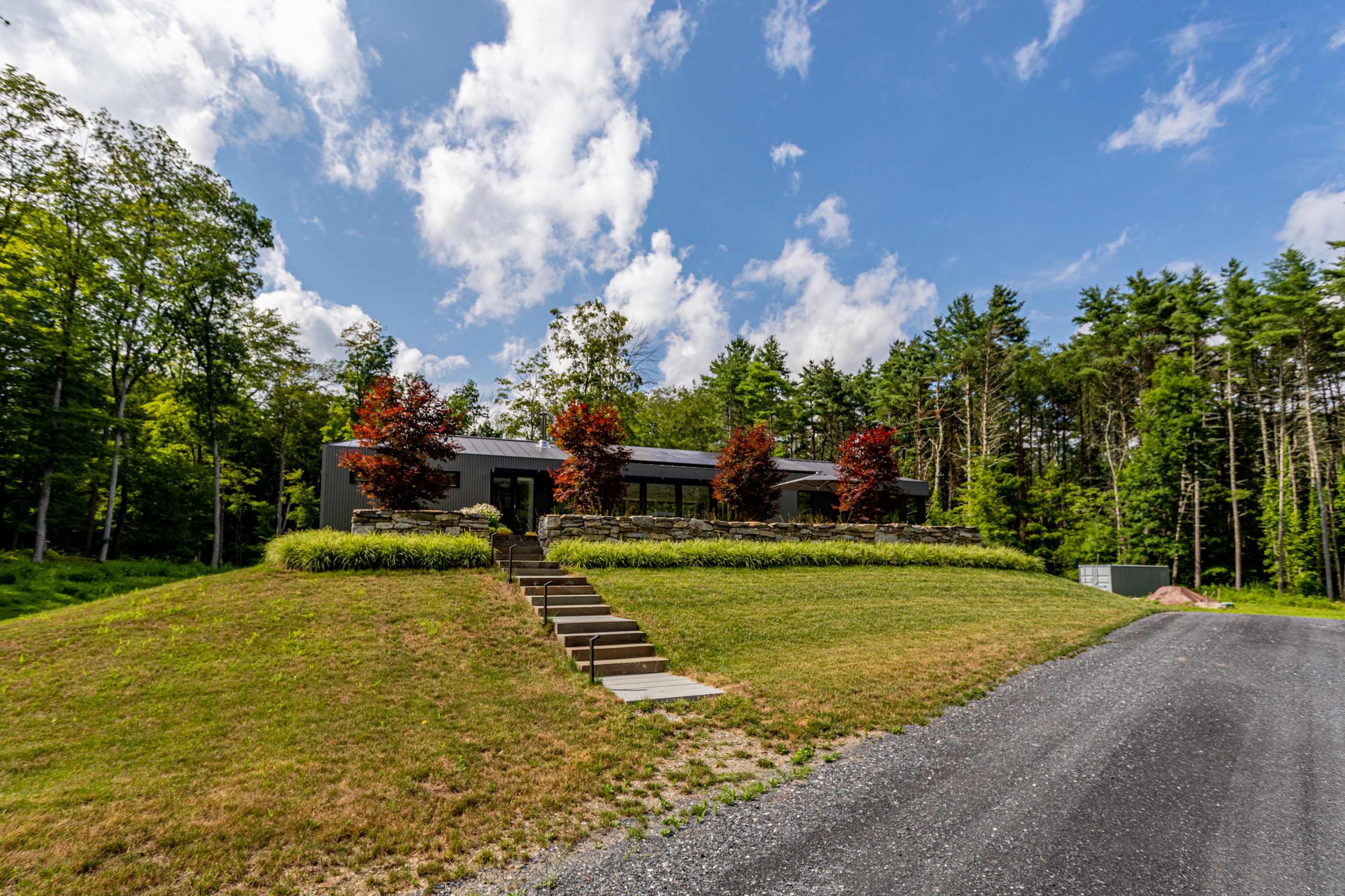The modern dark-colored house, featuring large windows and a sleek design, is perched on a grassy hill with a stone retaining wall and steps leading to the entrance, framed by mature trees and a gravel driveway.