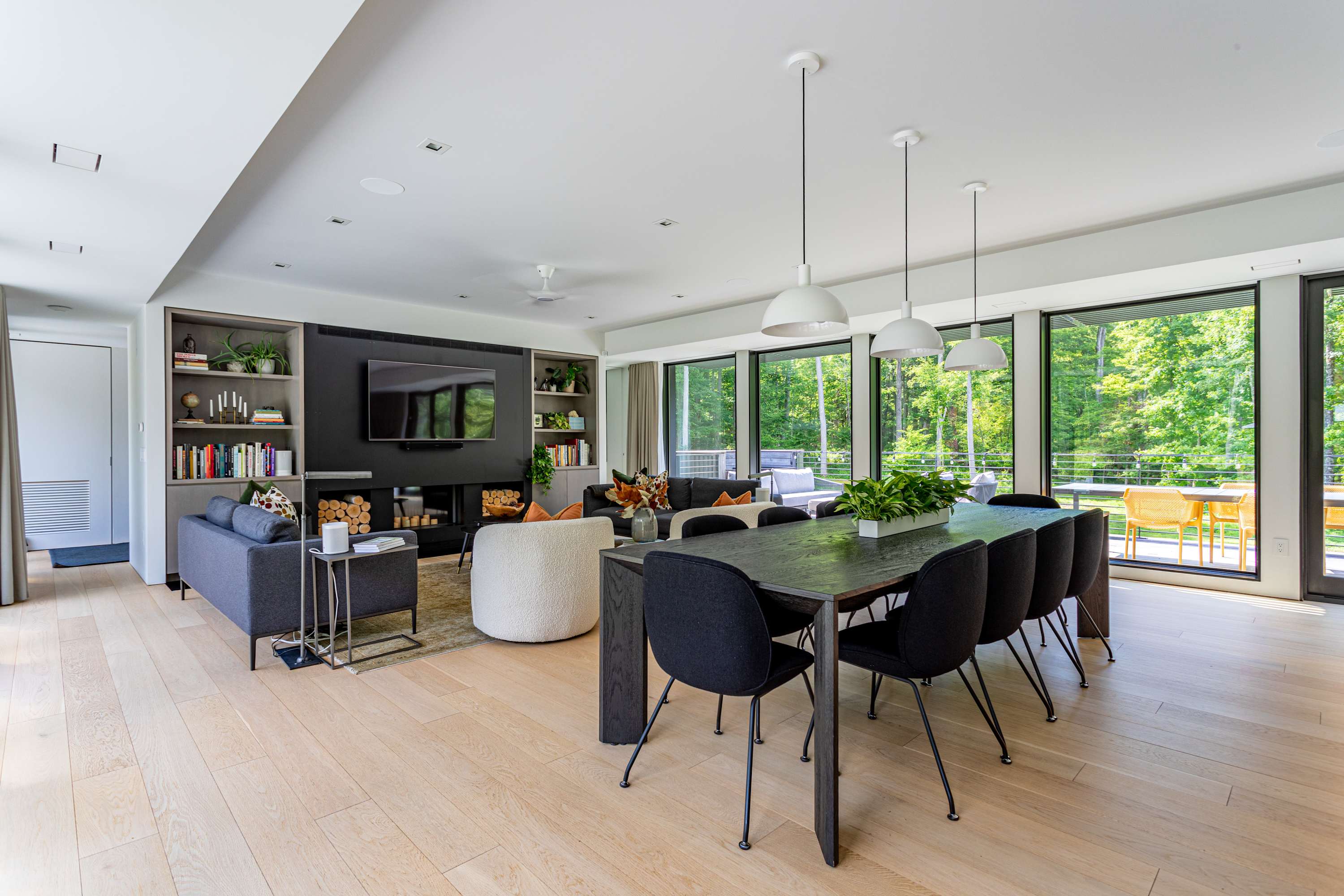 A modern open-concept living and dining space featuring a long dark wood dining table with black chairs, light hardwood floors, and a living area with a dark accent wall, built-in shelving, a TV, and a fireplace. Large windows offer views of lush greenery.