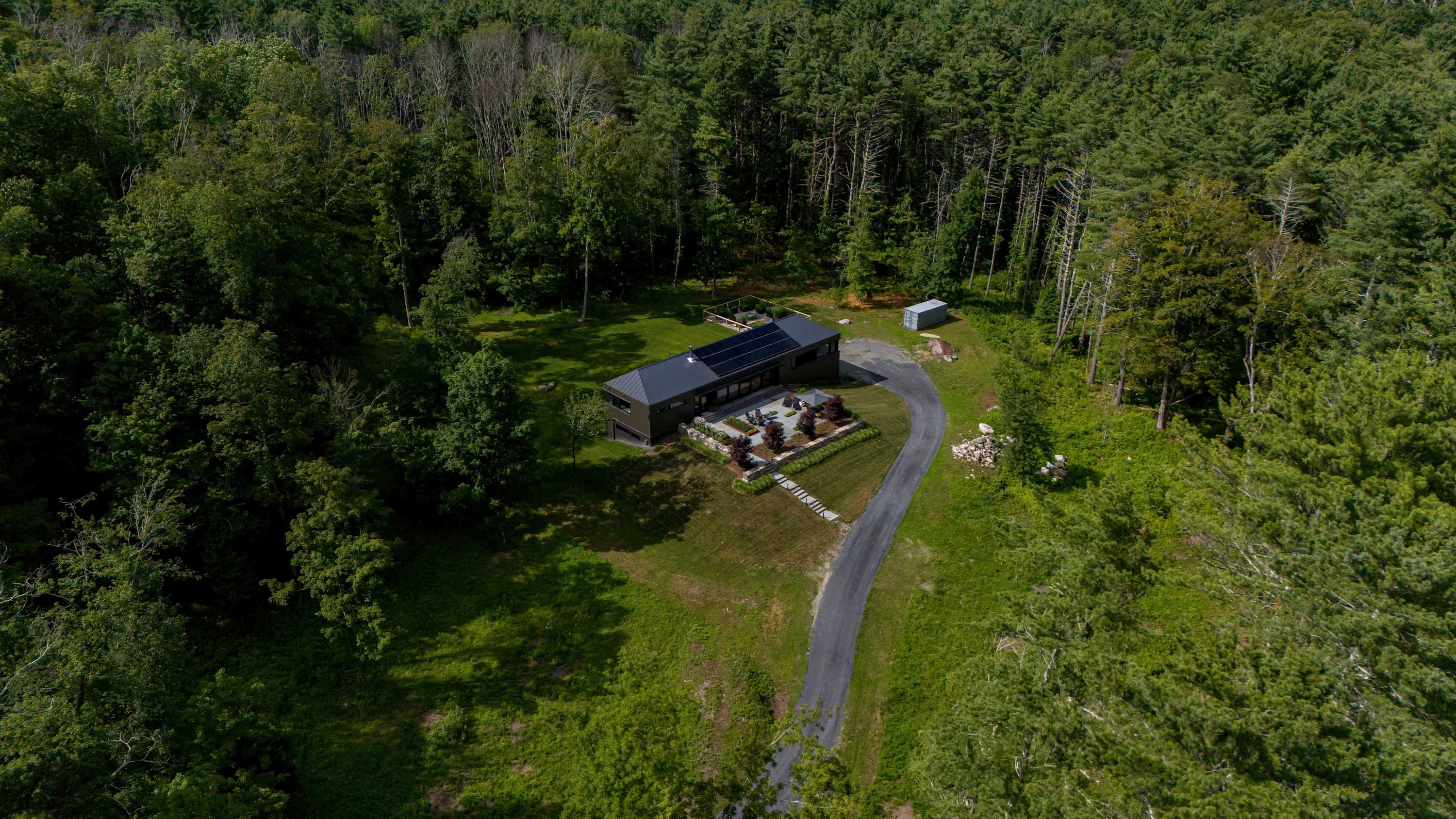 Aerial view of a modern house with a dark roof and solar panels, nestled within a dense green forest, featuring a long driveway and an expansive outdoor patio area, showcasing its secluded setting.