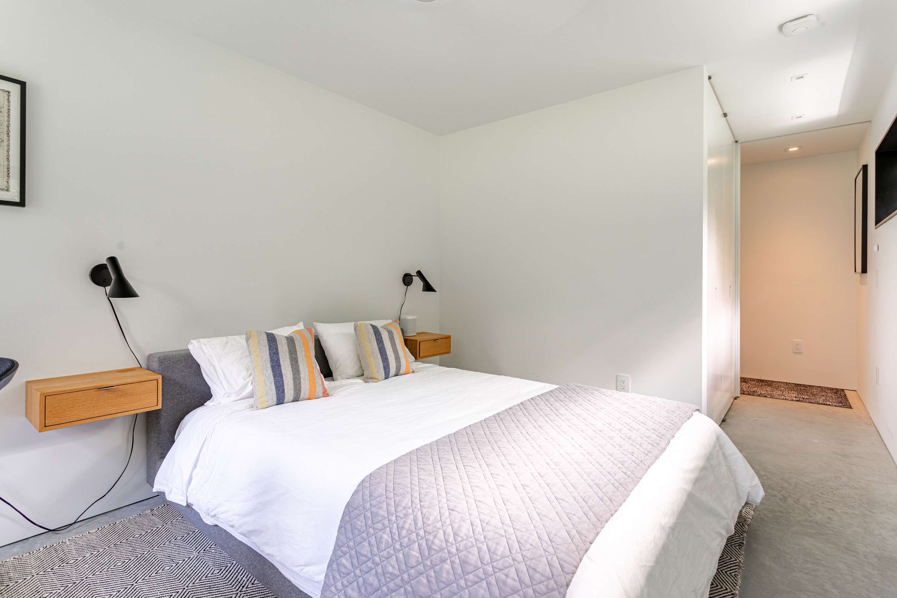 A closer view of the modern bedroom featuring a white queen-sized bed with a grey quilted throw and striped pillows. Floating wooden nightstands with black wall-mounted lamps flank the bed, and a patterned rug is visible on the concrete floor.