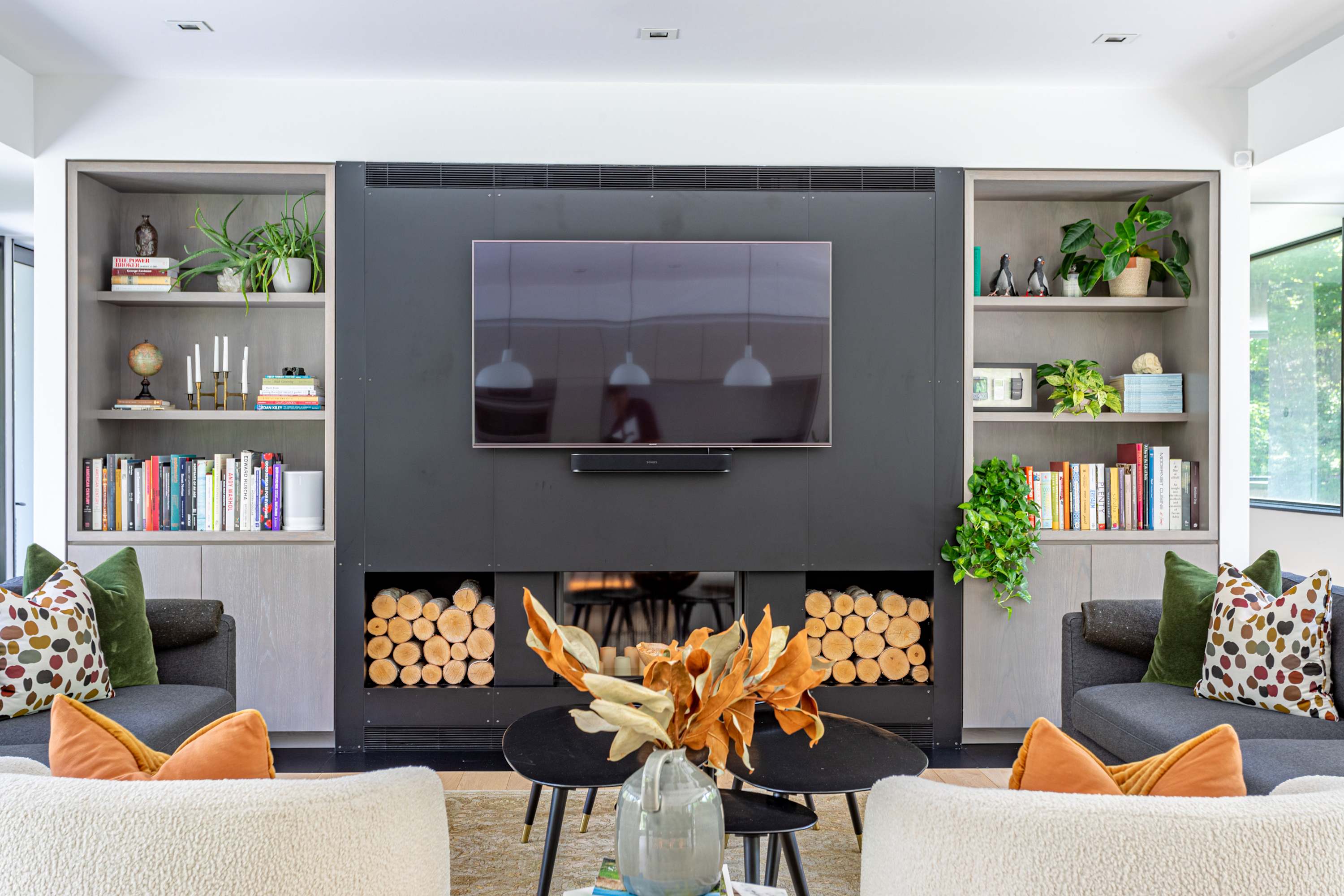 Contemporary living room with a large wall-mounted TV above a modern fireplace, flanked by built-in bookshelves filled with books and decor, and two dark sofas facing each other with colorful throw pillows.