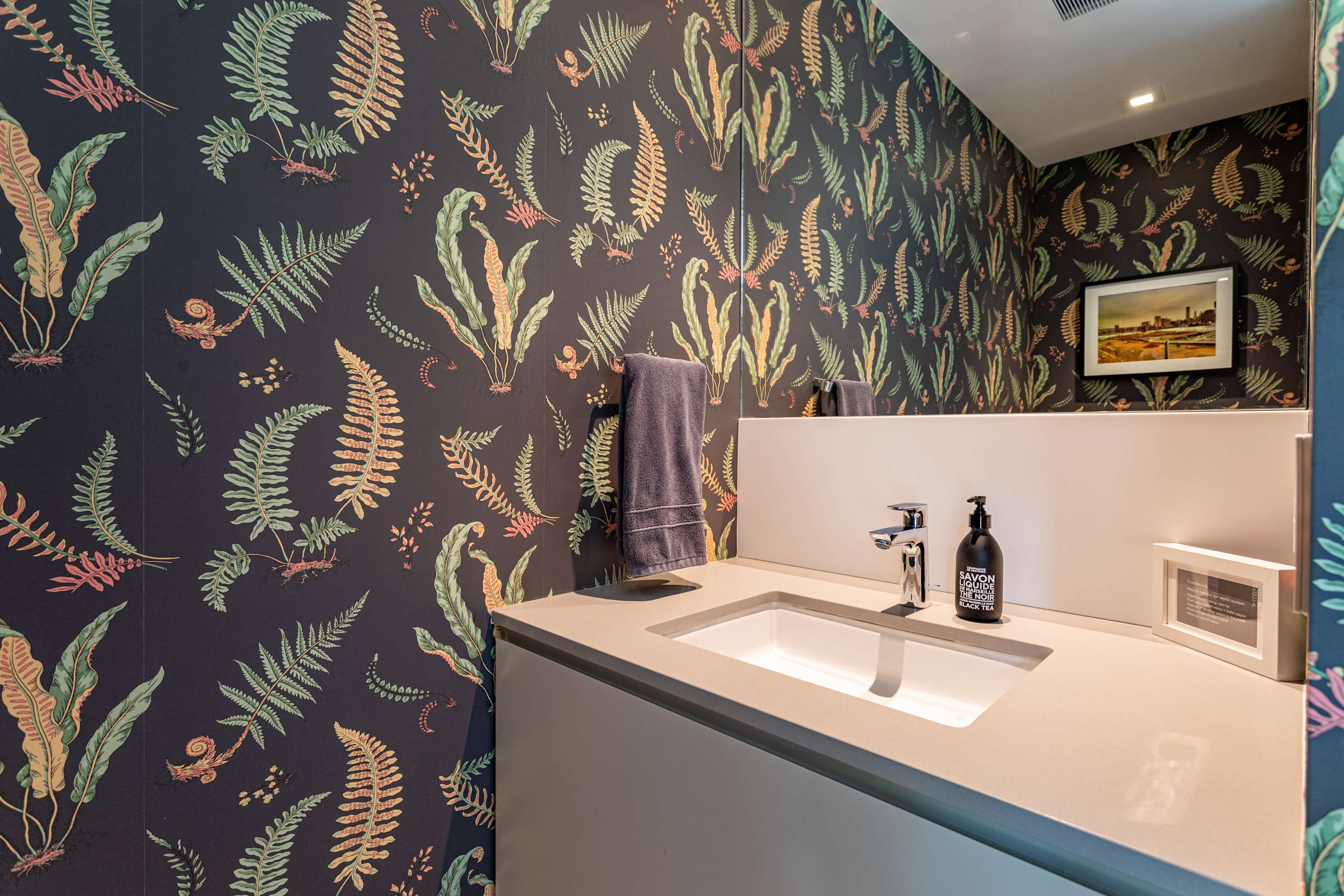 Stylish powder room with dark botanical wallpaper featuring green and orange fern patterns. A modern floating vanity with a white sink and a large mirror is present, along with a black soap dispenser.