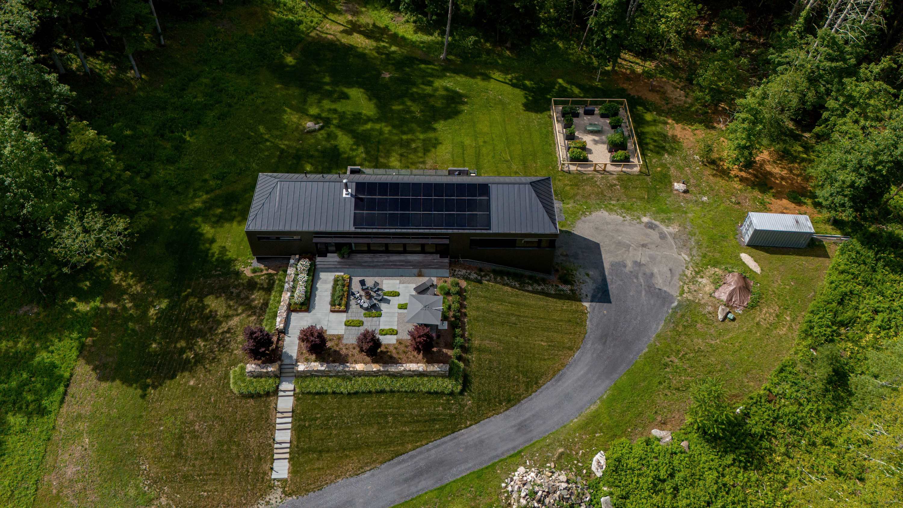 An overhead view reveals a modern house with a dark metal roof and solar panels, a spacious patio with outdoor furniture, and a gravel driveway, all nestled within a clearing surrounded by dense forest, with a separate garden area visible.