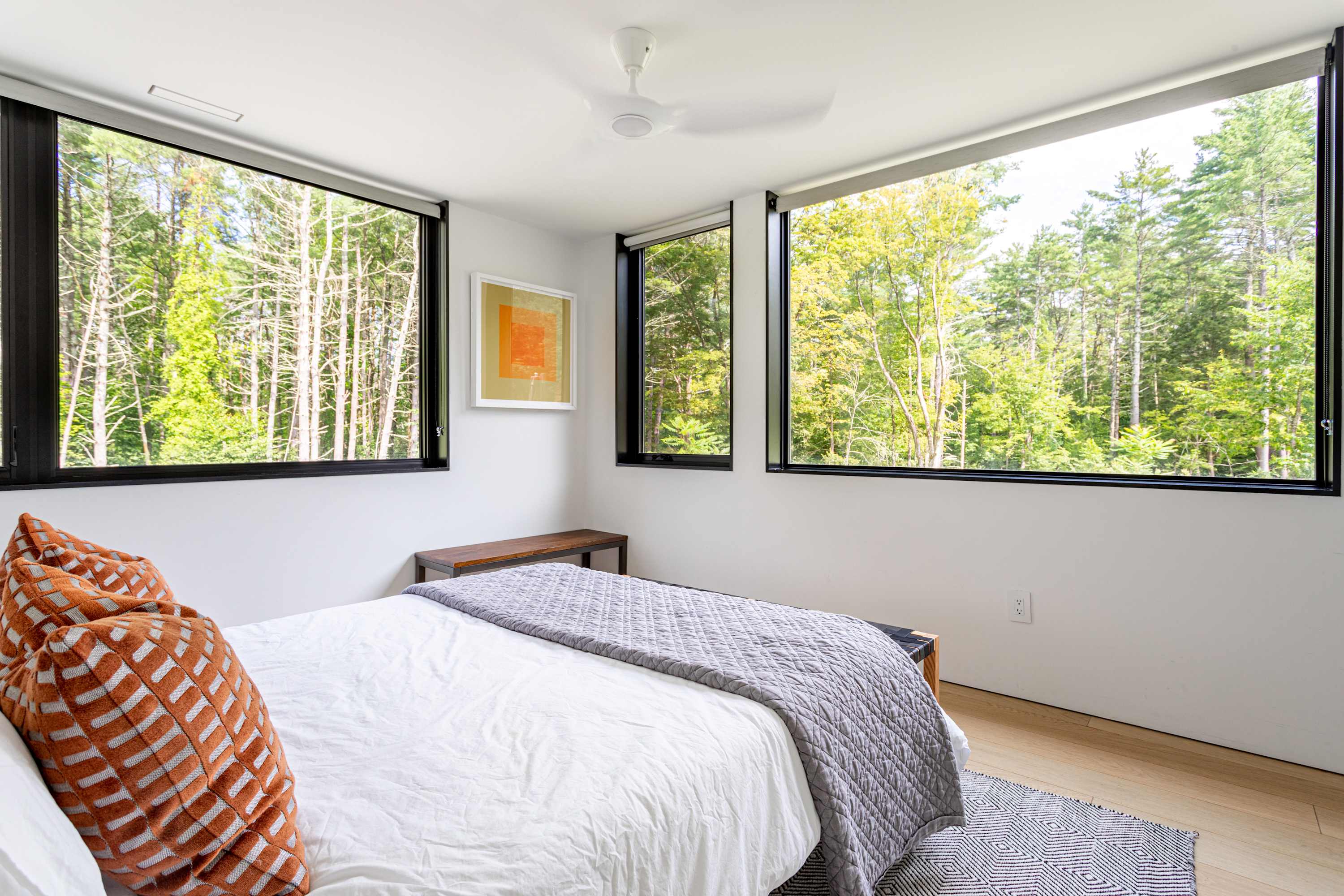 A bright bedroom-2 showcasing multiple large windows that provide panoramic views of a dense forest. The room has light hardwood floors, a queen bed with white and grey bedding, and a black and white patterned rug.