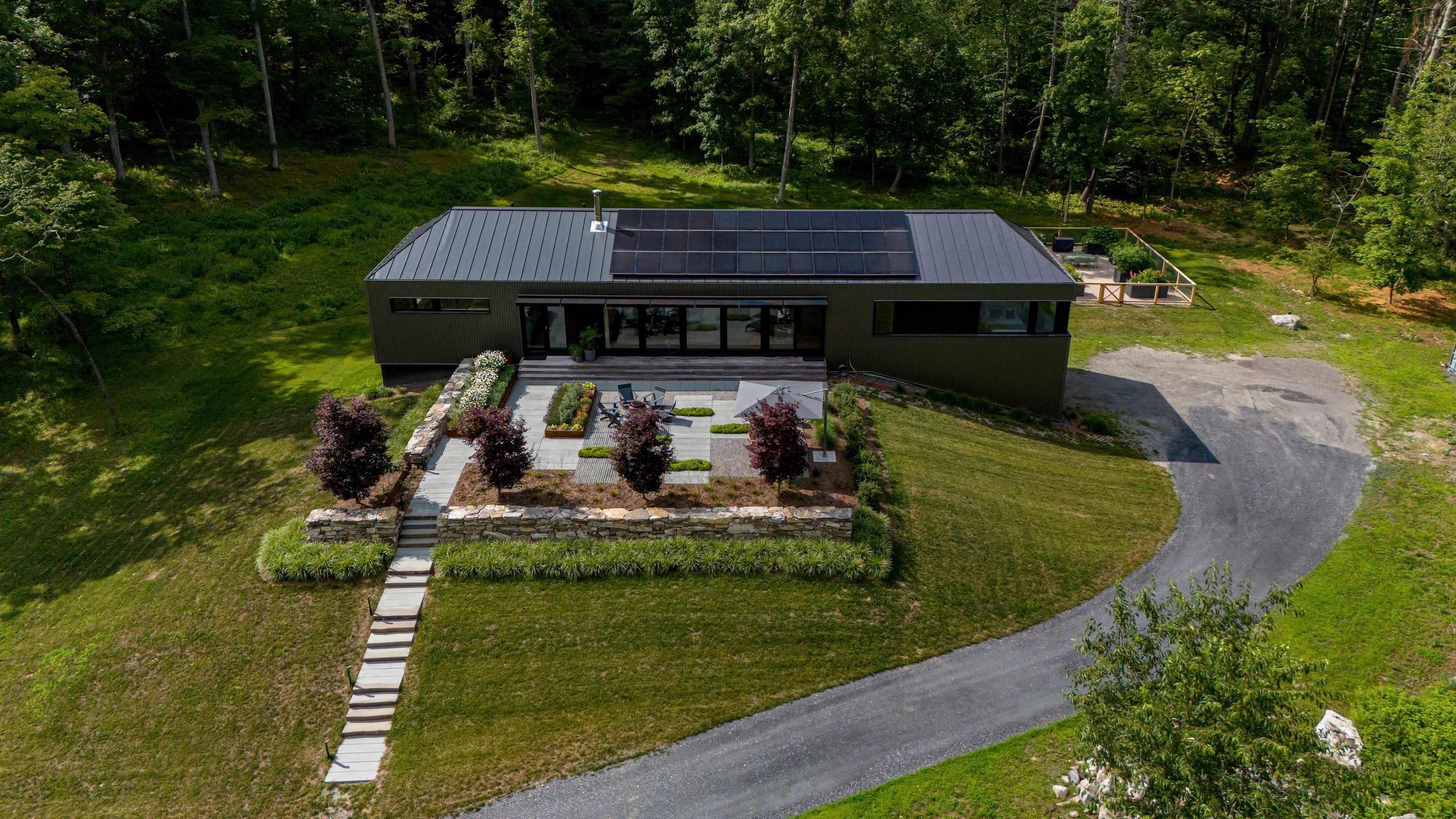 This aerial perspective showcases a contemporary dark-colored house with a metal roof and solar panels, complemented by a spacious stone patio featuring outdoor seating and decorative trees, bordered by a stone wall and a lush green lawn.