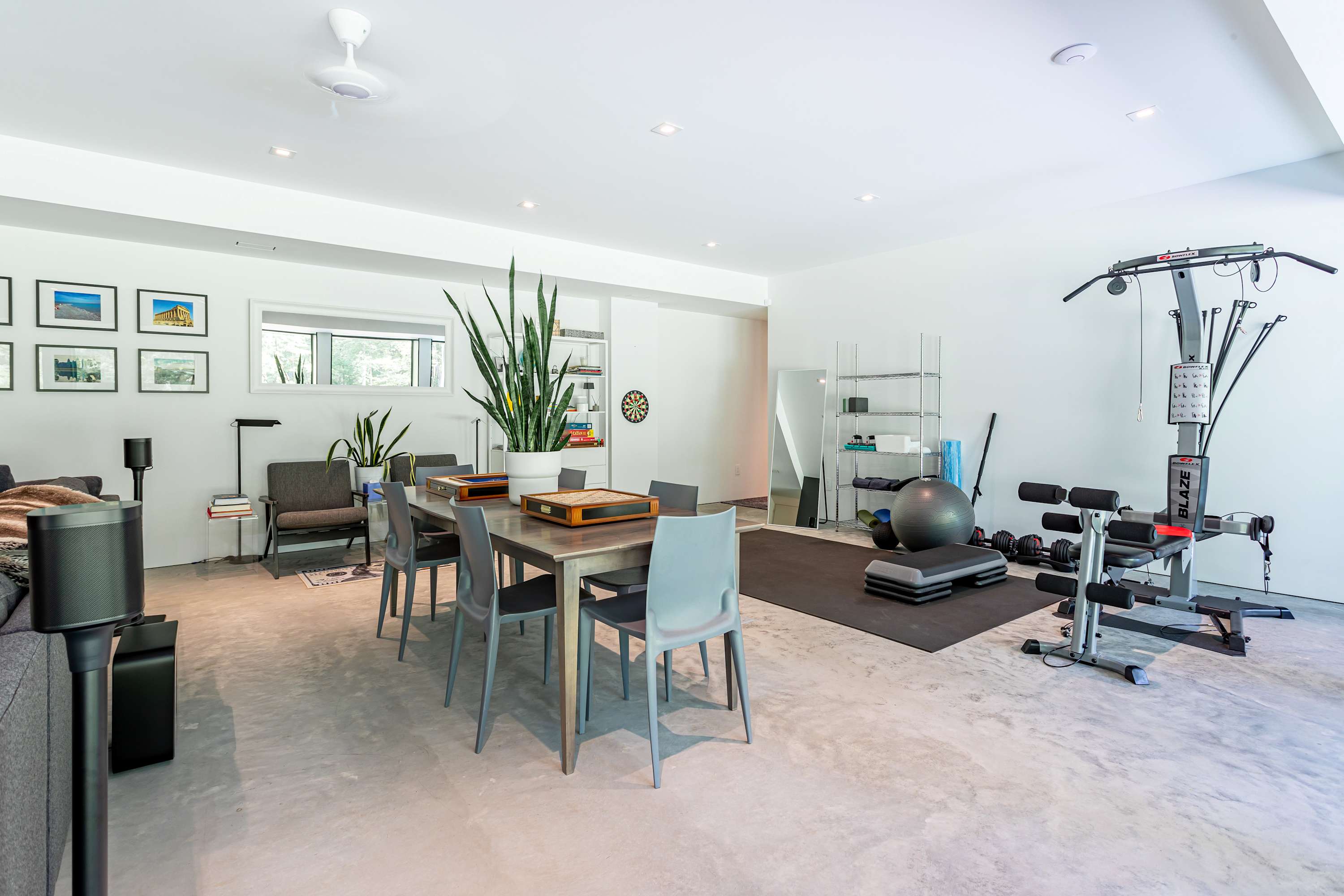 Open-concept recreational space featuring a long dining table with grey chairs, suitable for games, and a dedicated home gym area with various exercise equipment on a black mat. Large windows provide natural light and views.